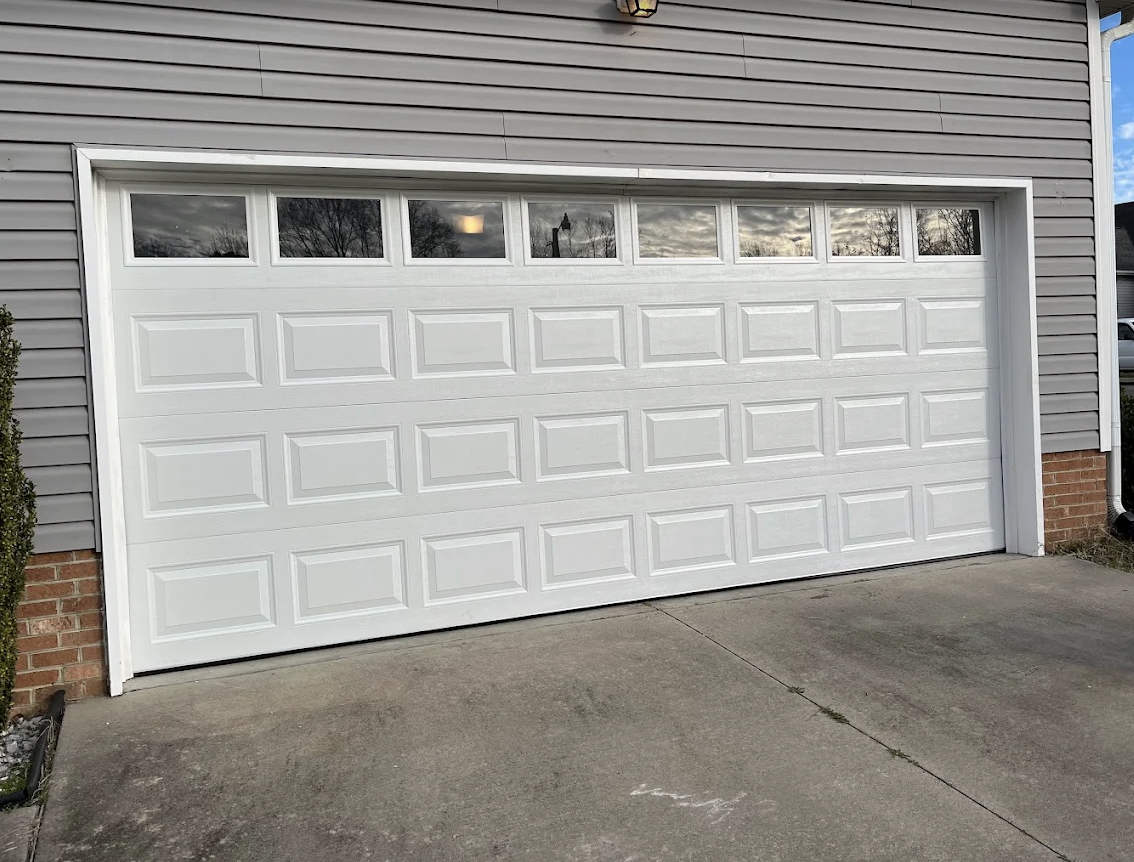White sectional garage door with rectangular panels and small glass windows at the top, installed in a house with gray siding and brick accents.