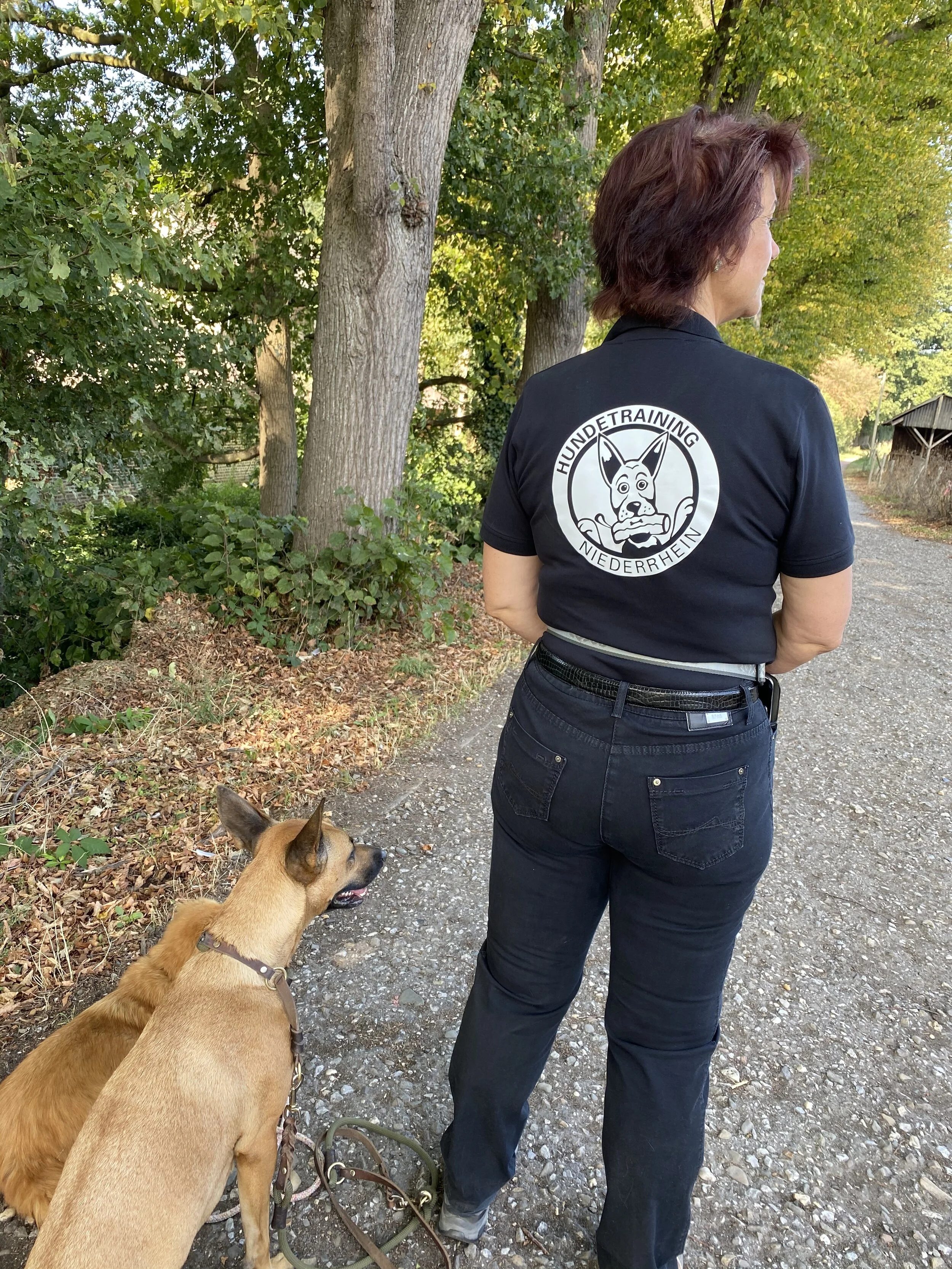 Eine Frau trägt ein schwarzes T-Shirt mit einem logo des Hundetrainings, die auf einem Weg in der Natur steht, neben einem braunen Hund mit Schleppleine.