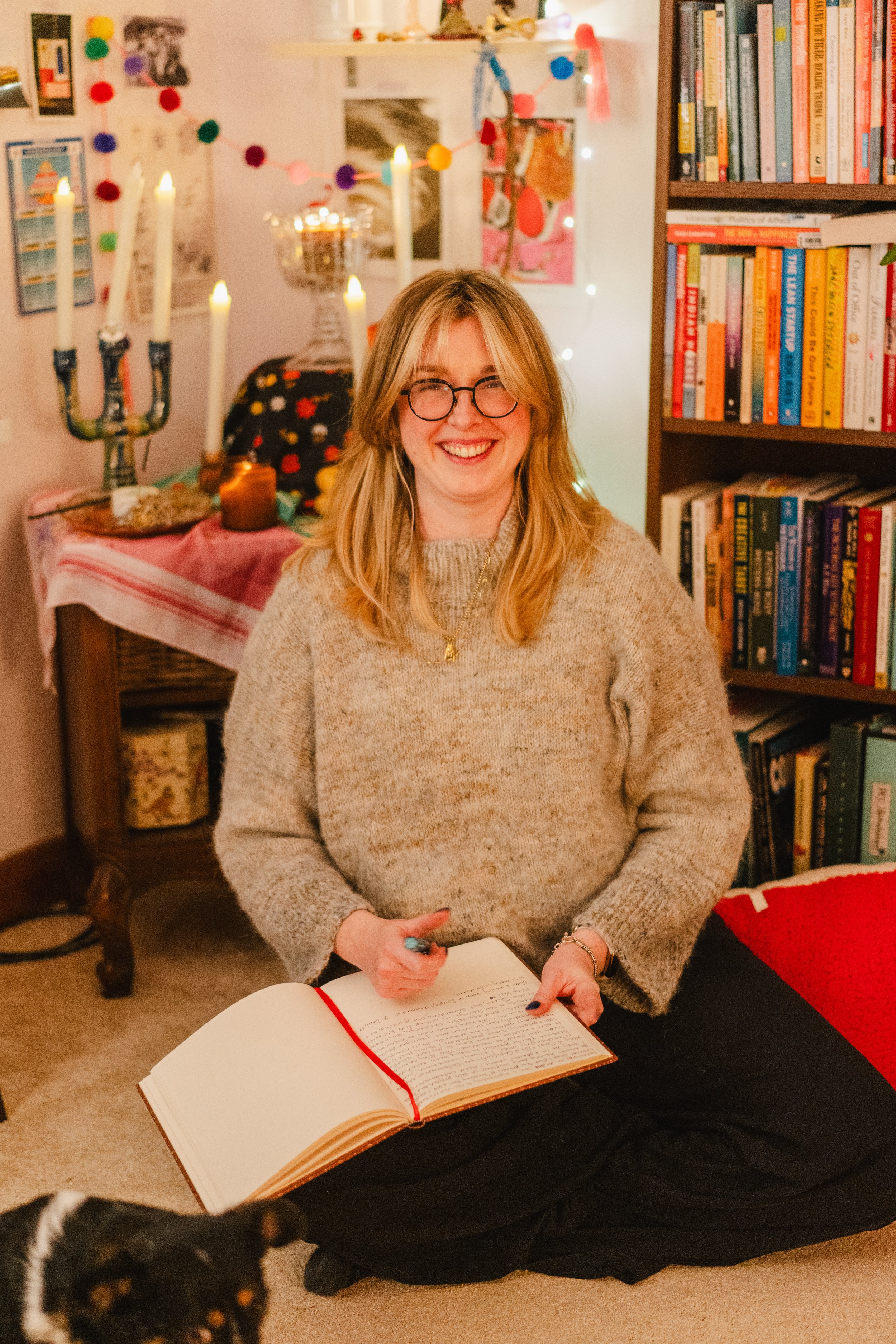 a woman sits on the floor journaling and smiling at the camera. there is a dog in the frame to the lower left