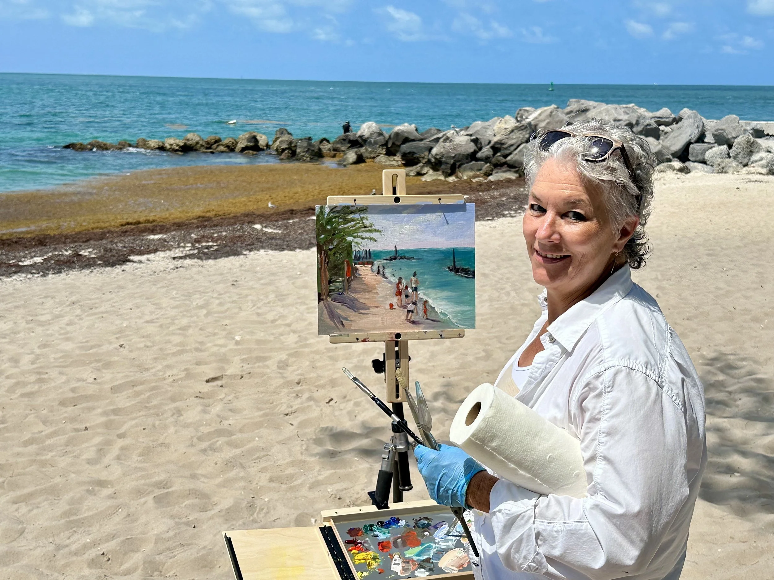 A woman with gray hair, wearing sunglasses on her head, painting outdoors on a sunny beach. She has a palette of colors and is painting a seaside scene with boats and water on a canvas mounted on an easel. The beach has sand, rocks, and the ocean with a clear blue sky above.