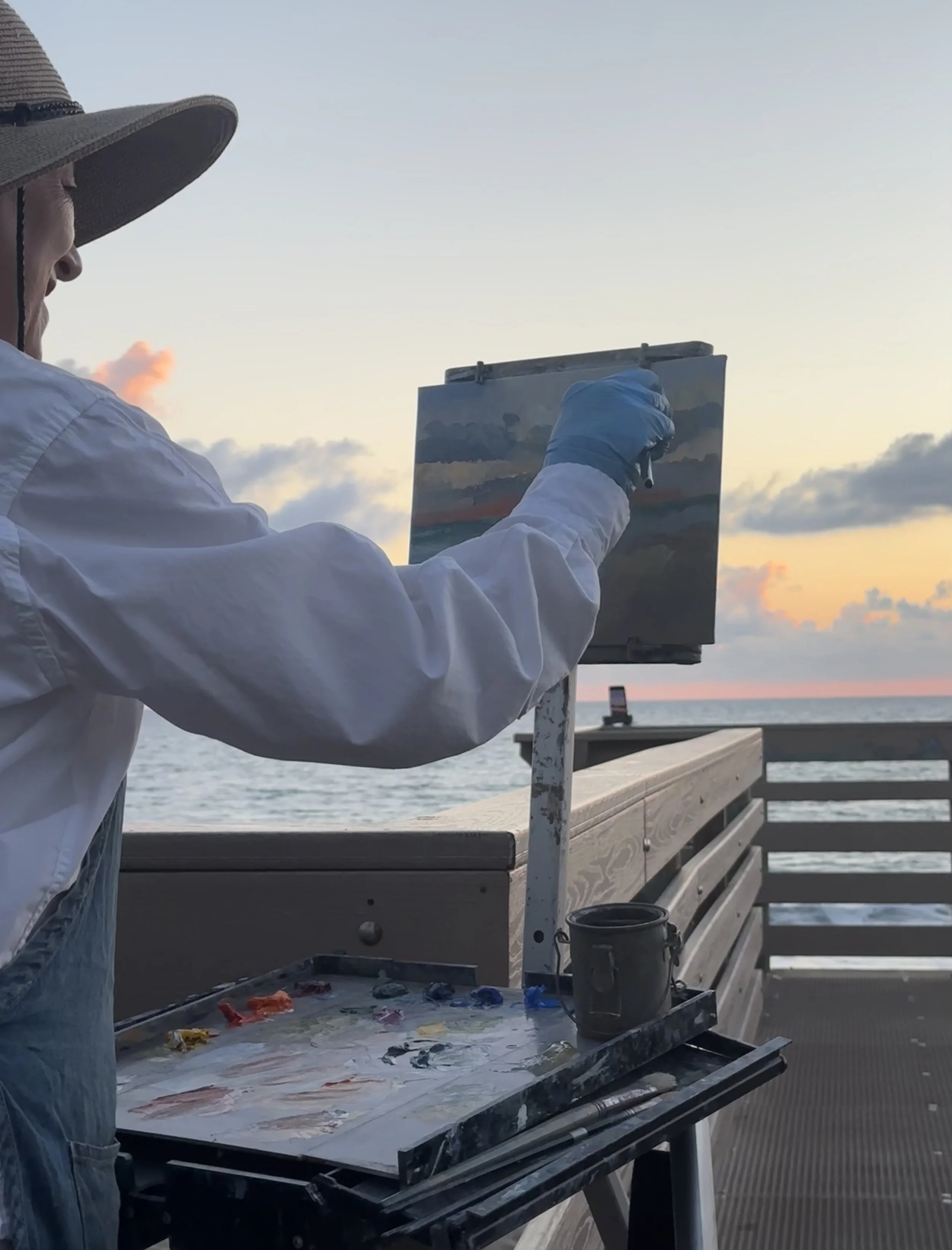 An artist painting outdoors on a boardwalk at sunset, with the ocean and cloudy sky in the background. The artist is wearing a wide-brimmed hat, a white long sleeve shirt, and a blue glove on their hand holding a paintbrush.