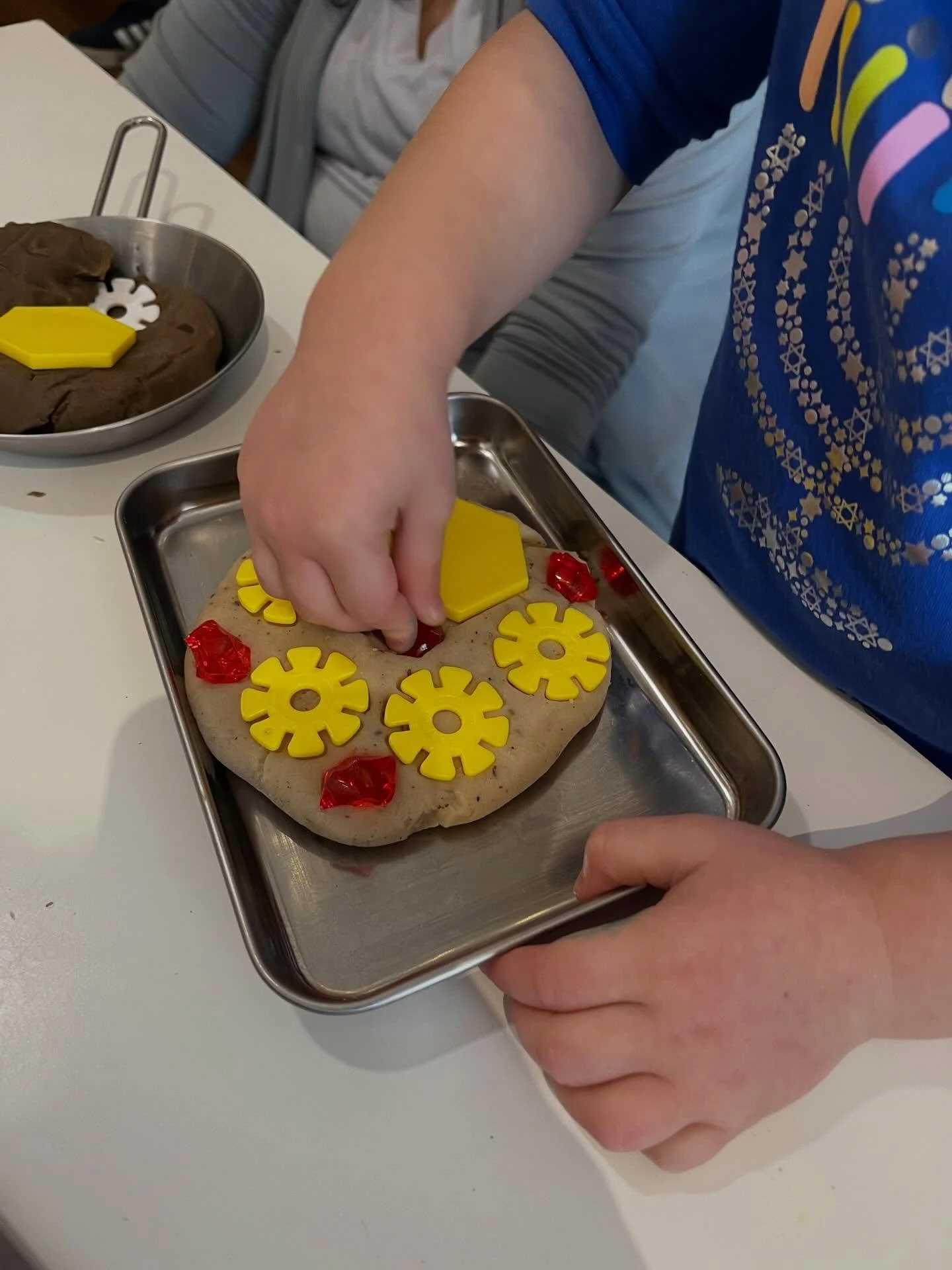 During our Hanukkah themed party, children pretended to fry up latkes and sufganiyot with play dough and loose parts. They used red gems as the jelly filling for sufganiyot, white loose parts as sour cream for latkes, and yellow loose parts as apples