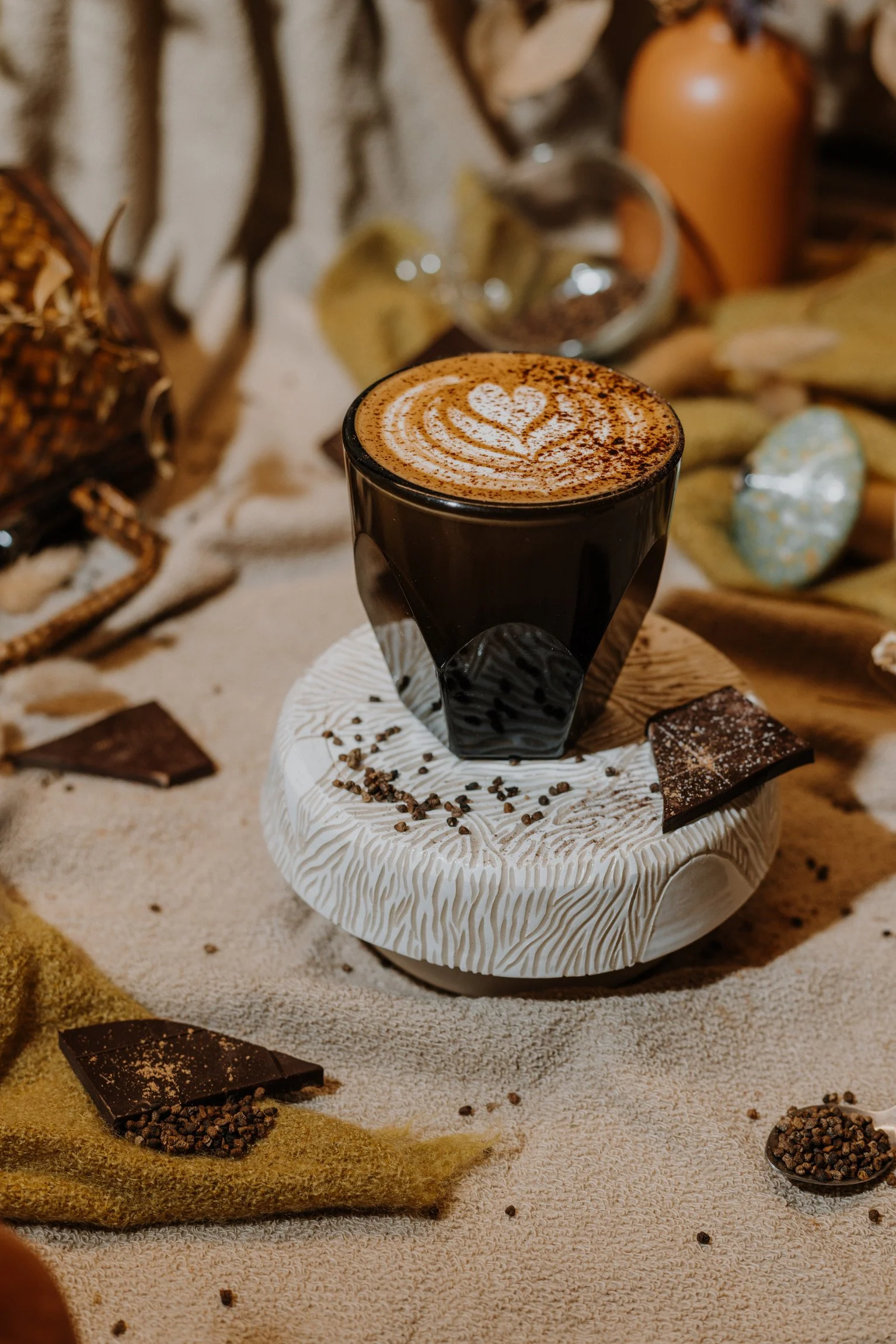 a dark glass cappuccino cup with latte art sits on a white ceramic pedestal, surrounded by chocolate bar pieces and cardamom seeds. 