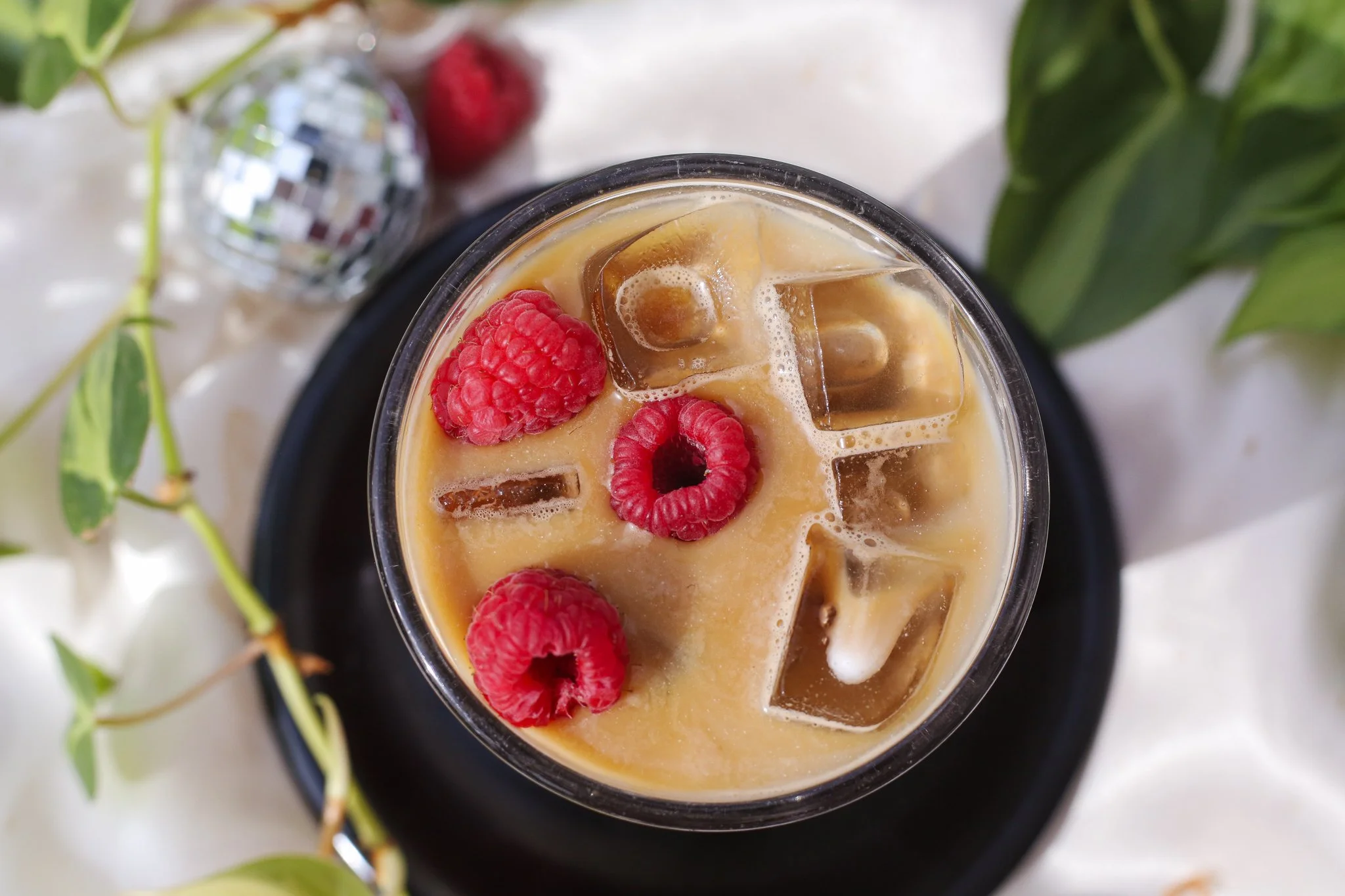 top-down view of an iced latte with fresh raspberries floating on top. we can see leaves and a disco ball and white fabric in background..