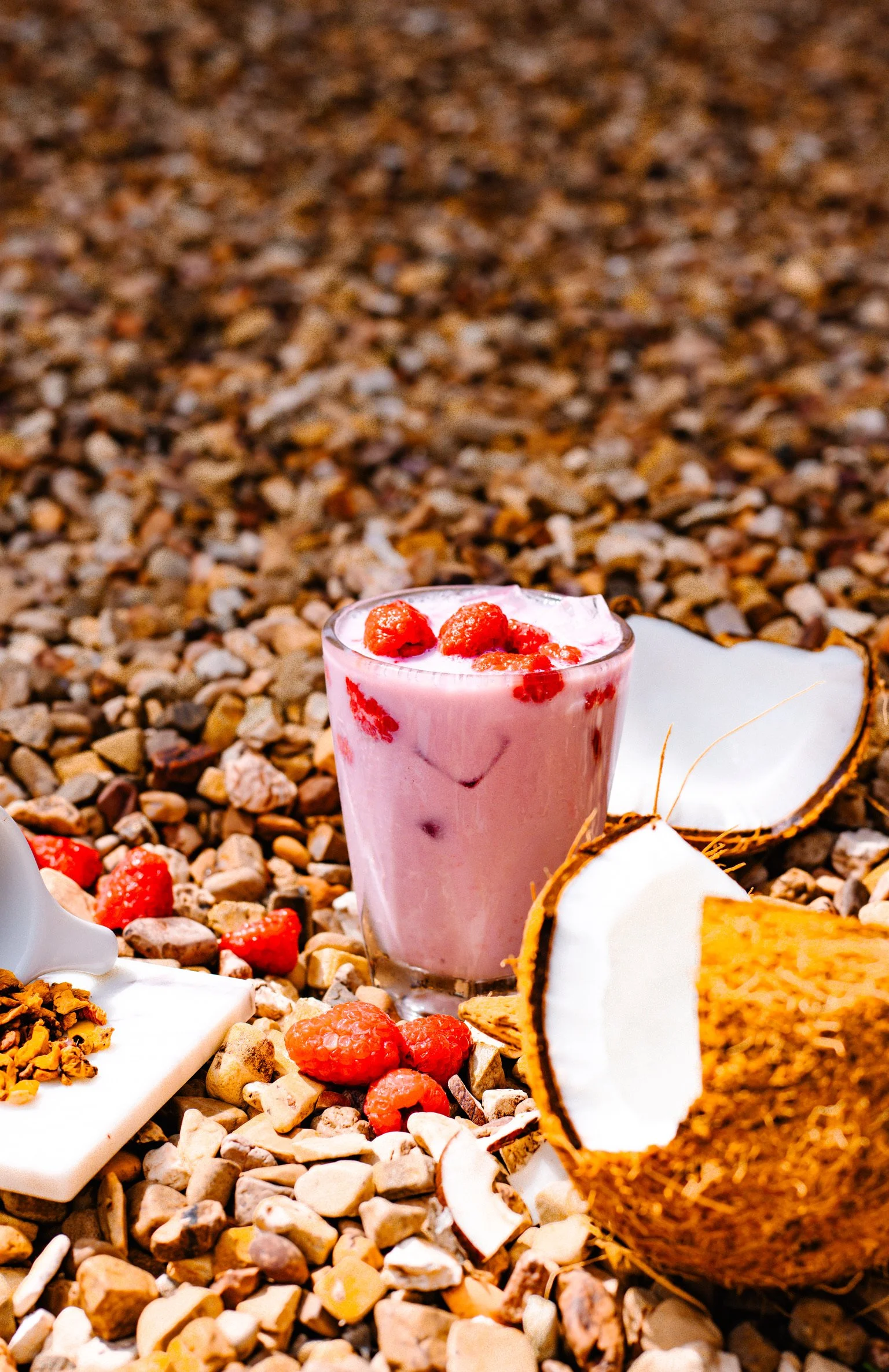 a glass filled with an iced pink drink topped with fresh raspberries sits on a bed of rocks, surrounded by raspberries and pieces of coconut