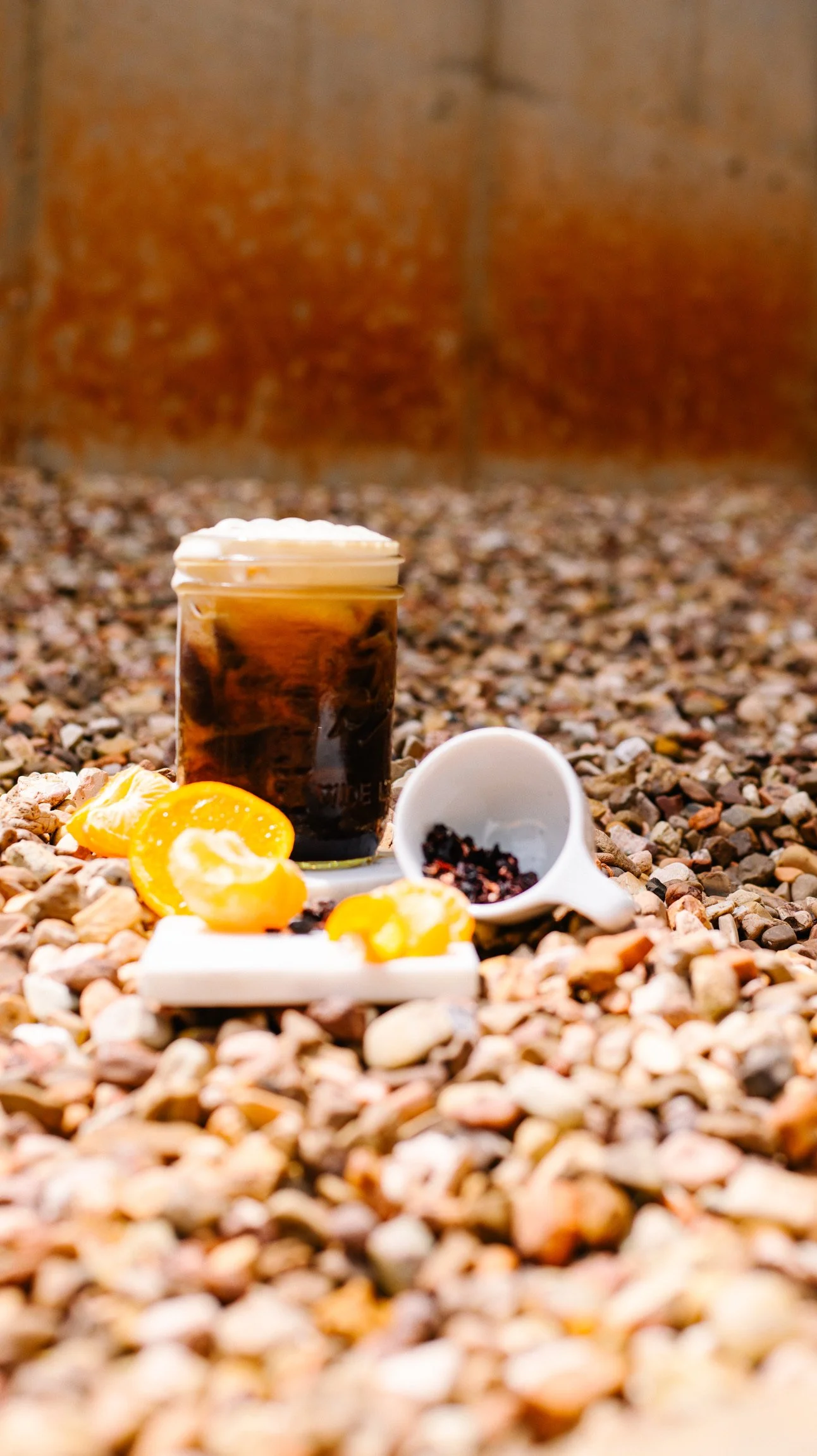 a mason jar full of iced coffee is topped with overflowing foam. the jar is surrounds by rocks. orange slices and loose leaf tea sit in front of the drink.