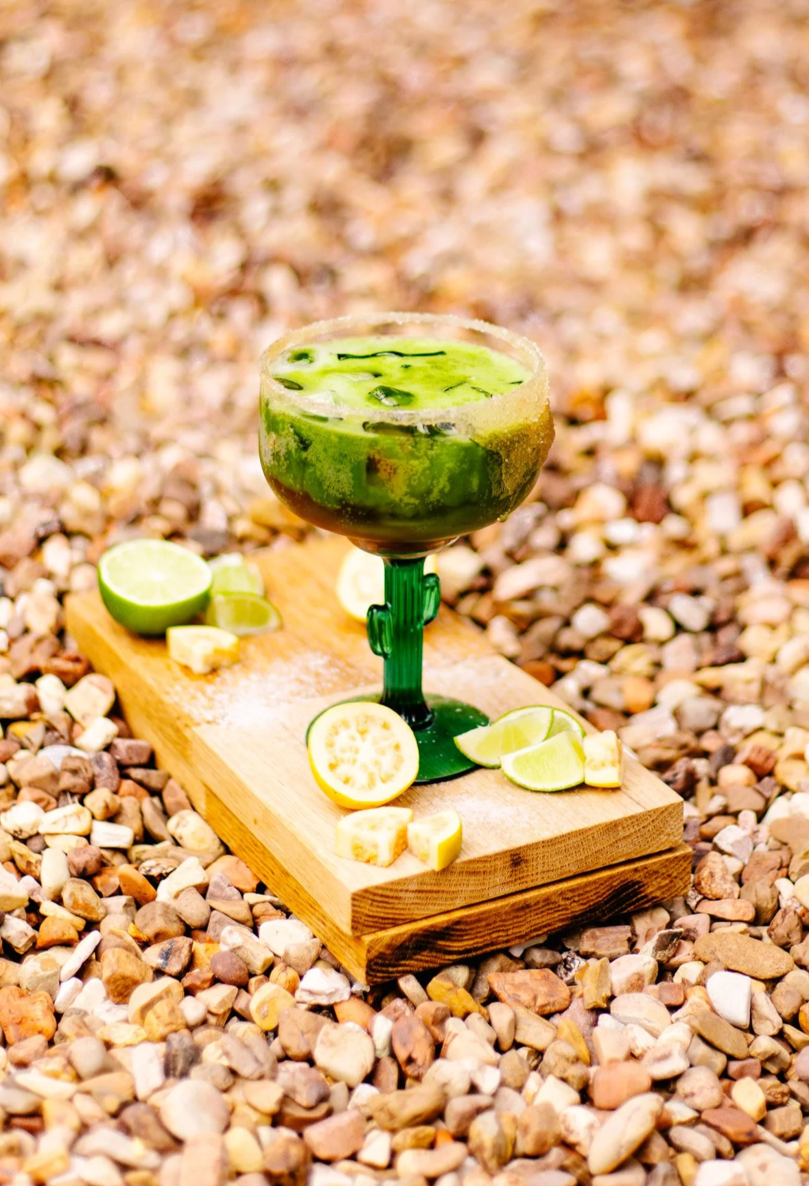 a deep bowl, tall green stem glass has a salted rim and is filled with sparkling matcha drink. the glass sits on a wooden board that has sliced and halved limes and guava and salt on it. the drink is surrounded by rocks.