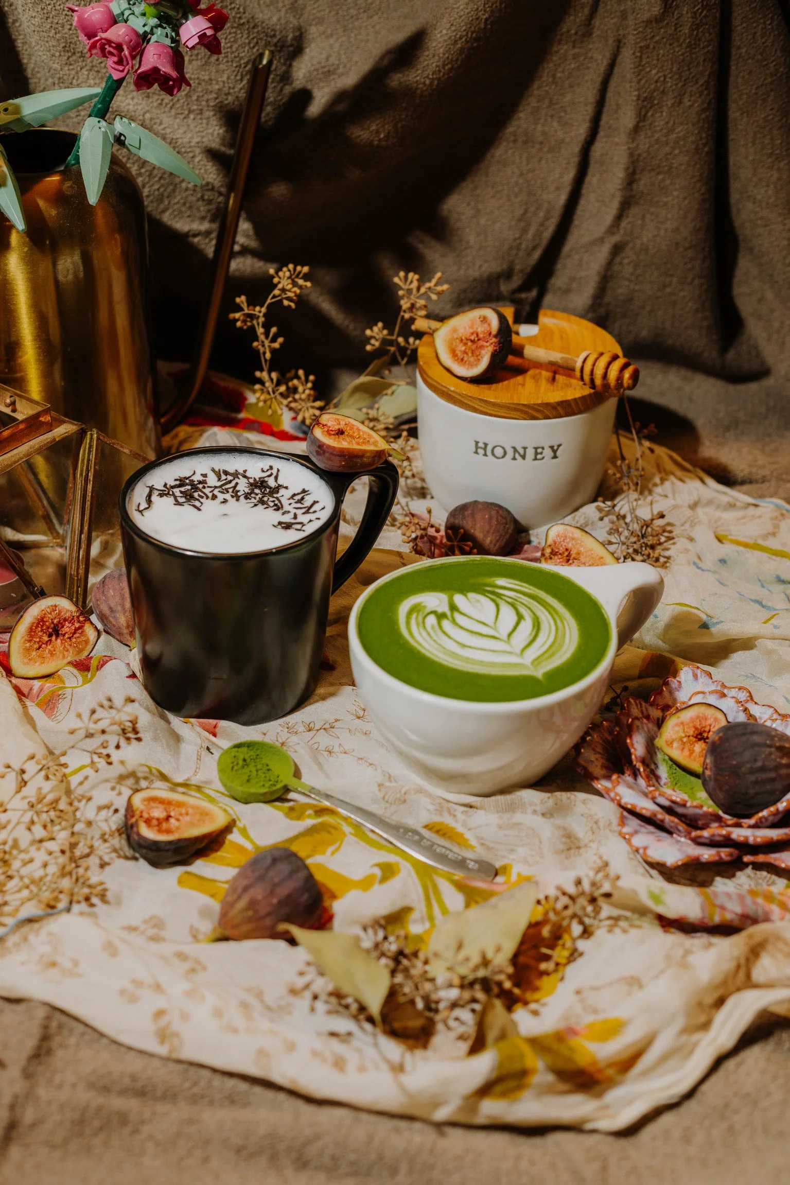two mugs sit on a fabric surface surrounded by figs and dried leaves. one mug is black and has a fig on the rim, filled with white foam. the other mug is white, filled with a matcha latte and latte art.