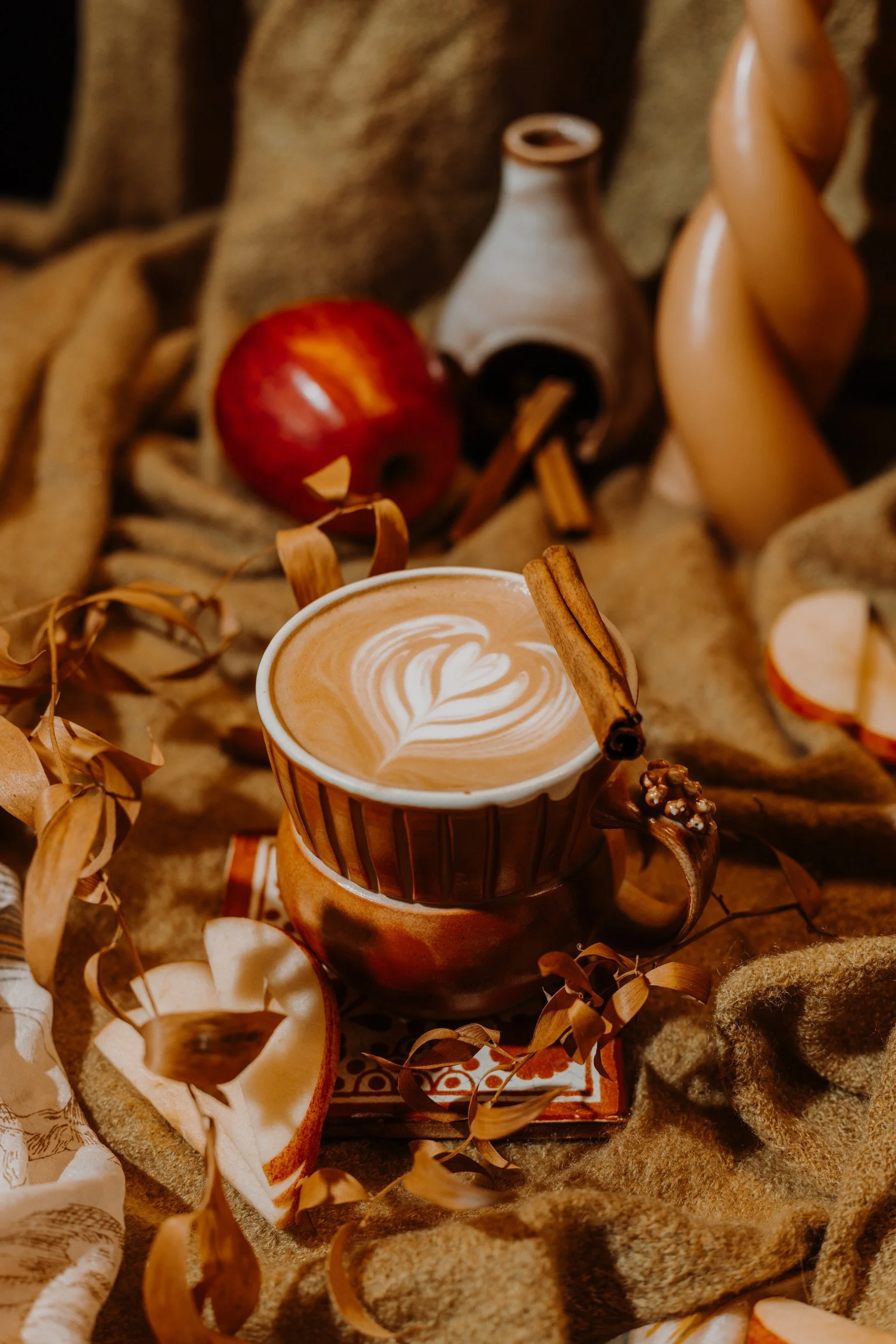 an orange mug sits on a fabric surface surrounded by dried leaves and sliced apples. there is latte art in the mug, and a cinnamon stick sits on the rim.