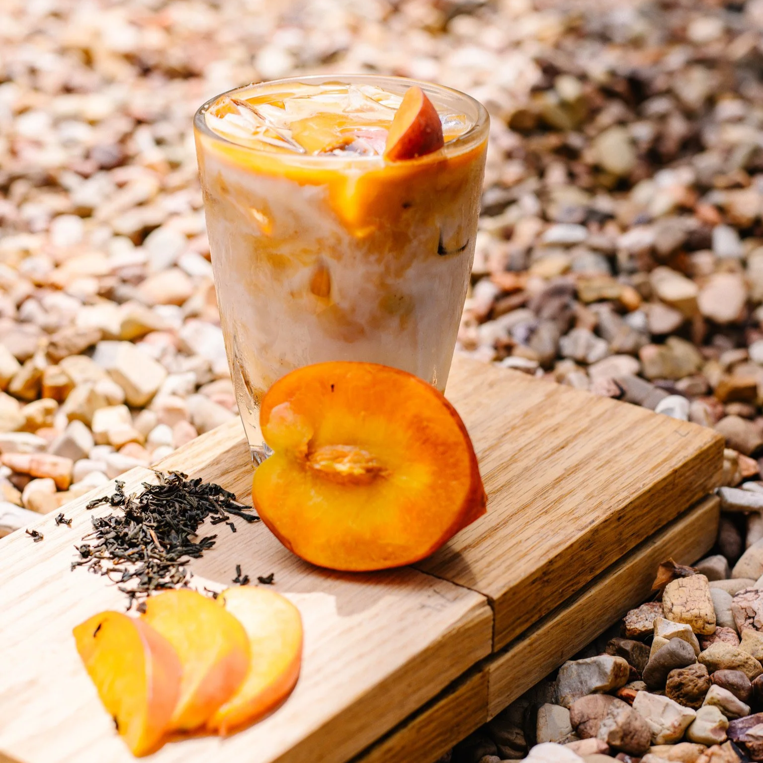 a glass filled with layered iced coffee sits atop a wooden surface that has sliced and halved peaches and loose leaf tea on it. the drink is surrounded by rocks.