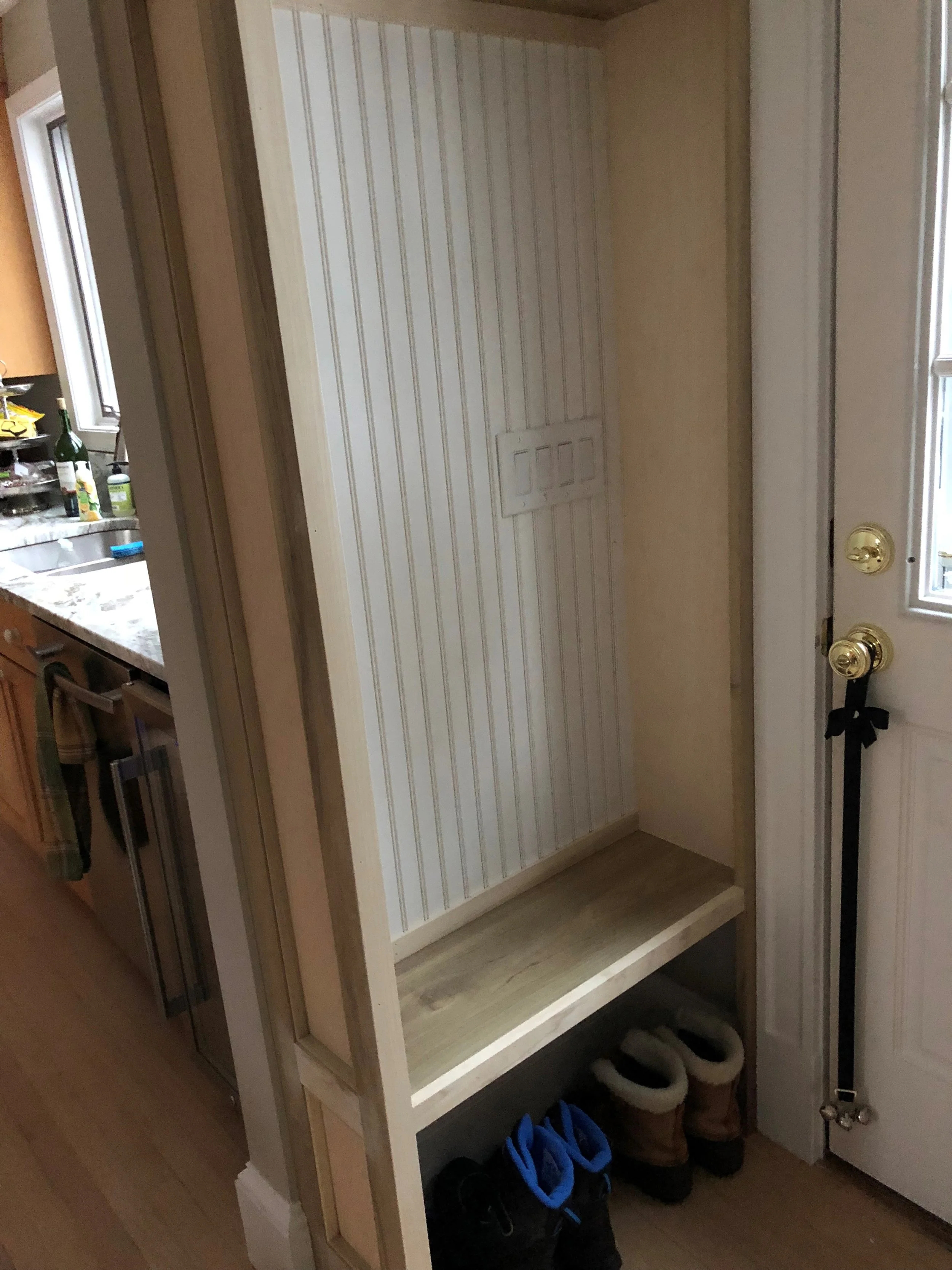 Interior of a house with a small shoe rack and shoes underneath. A beige striped wall with a small door panel is visible. Part of a kitchen is seen on the left with items on a kitchen counter.
