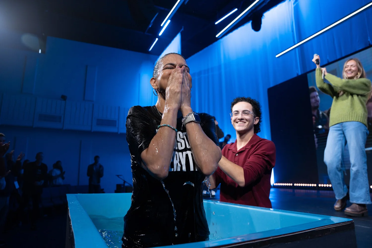 A woman standing waist-deep in a baptism tank with her hands over her face, water running off her, as a young man beside her claps and a woman on the stage behind cheers.