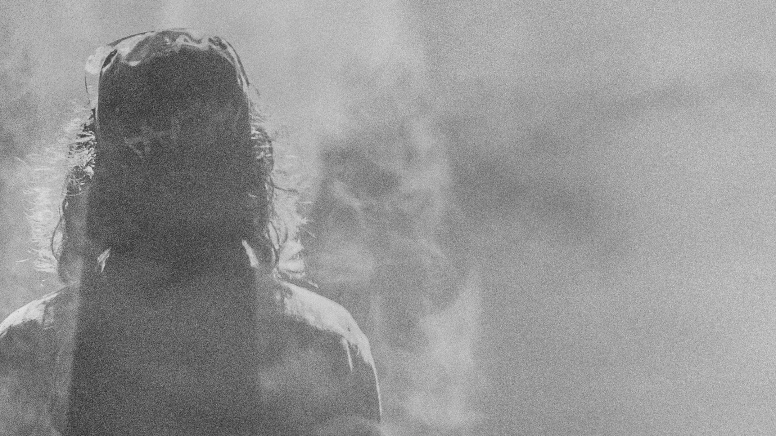 A black-and-white photograph, heavily grained, of a person seen from behind in thick mist, facing a second figure blurred by the fog ahead of them.