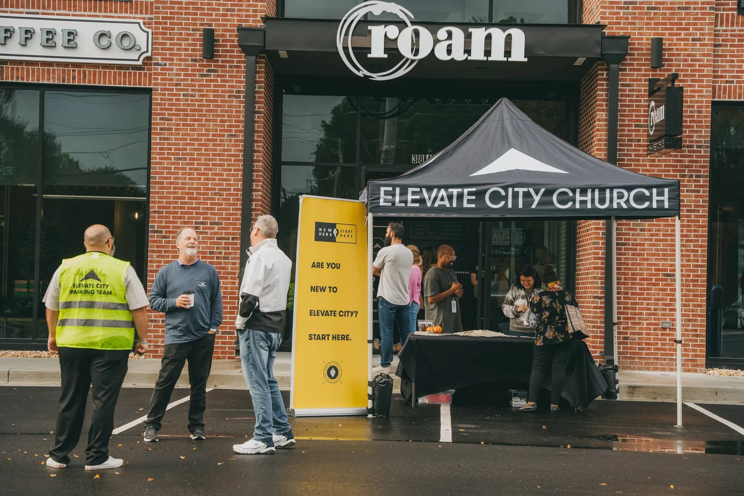 People arriving on a Sunday morning under the Elevate City Church tent at the entrance, a parking team volunteer in a hi-vis vest and a banner reading are you new to Elevate City, start here