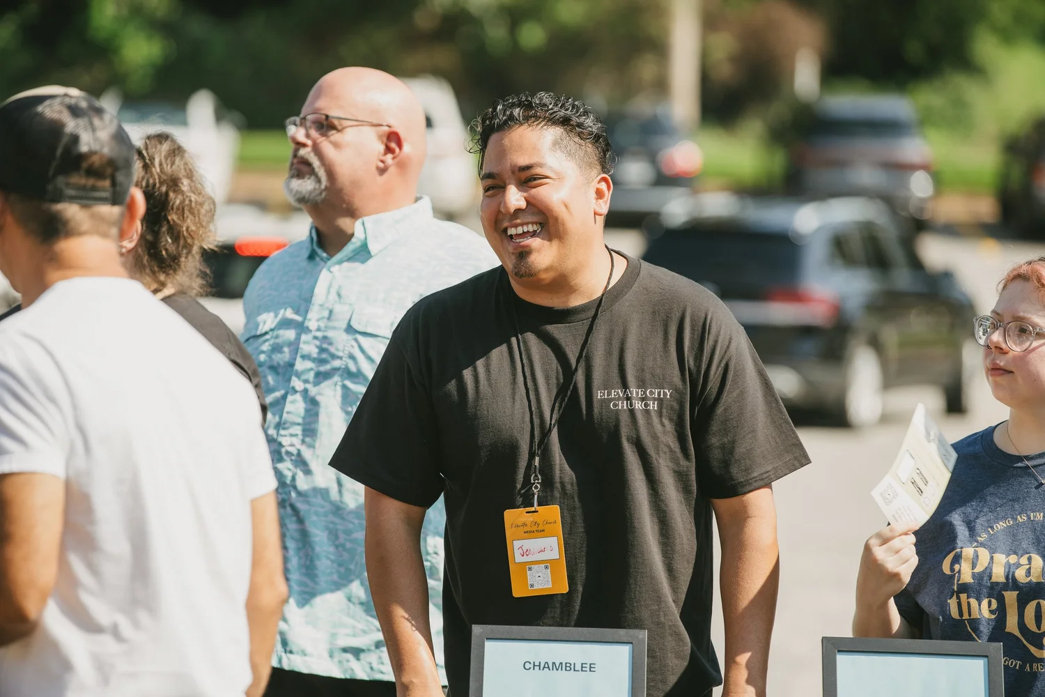 Outdoors on a bright day, a man in an Elevate City Church T-shirt and a media team lanyard laughing with a small group beside a stack of framed signs.