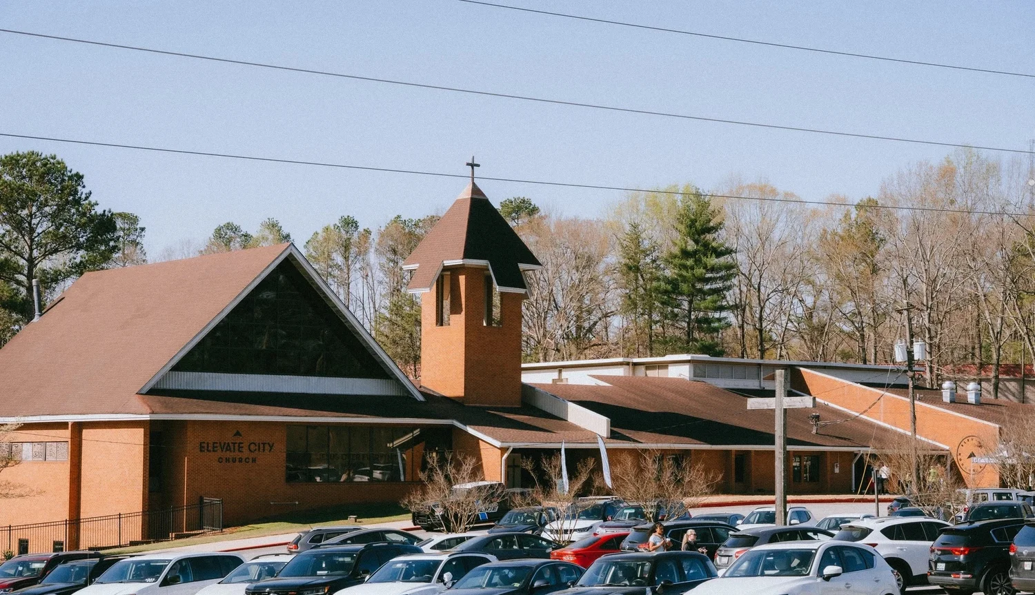 The Elevate City Church building at 7770 Roswell Rd: a brick A-frame with a bell tower and a cross, its car park full on a Sunday morning