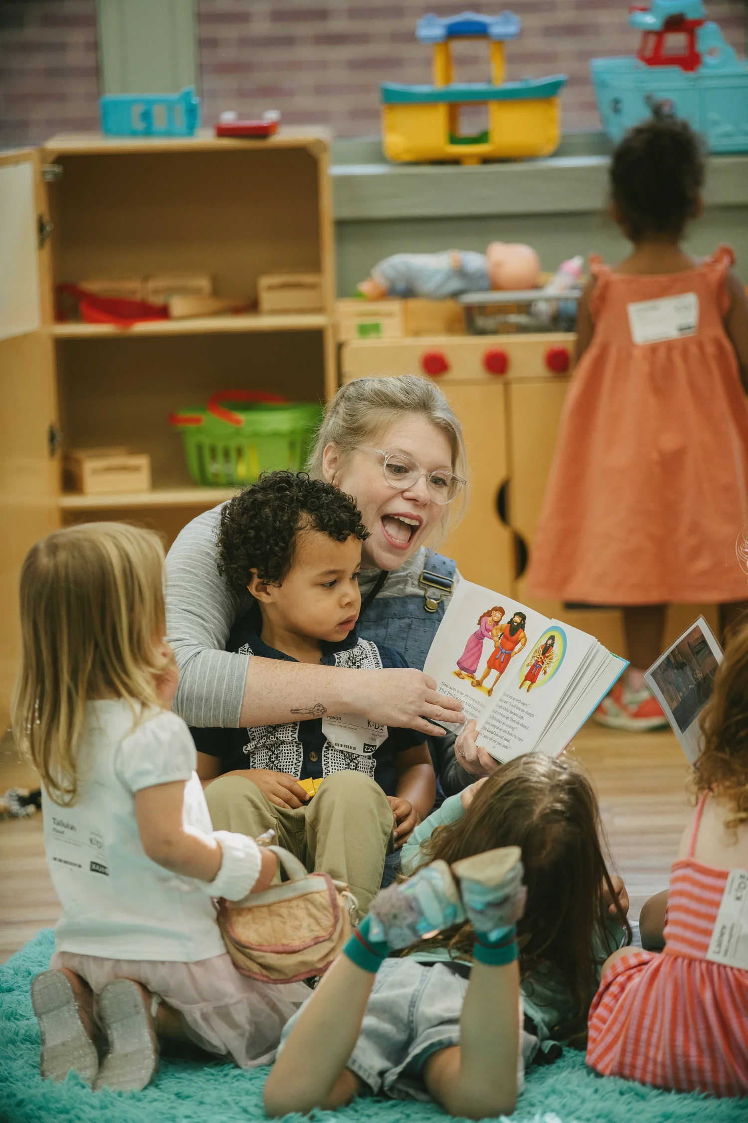 A woman sitting on a classroom rug reading an illustrated Bible storybook aloud to four small children gathered around her.