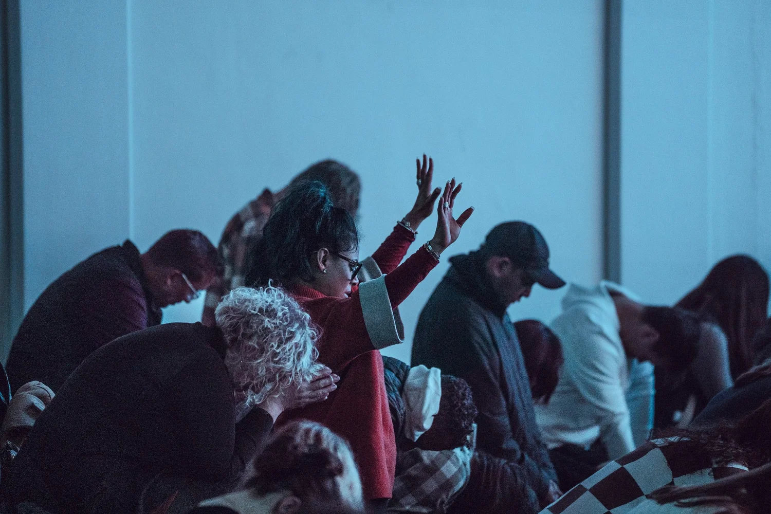 A room lit blue, rows of people bowed over their seats in prayer, one woman among them with a hand lifted.