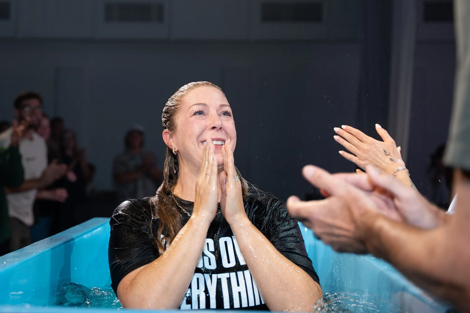 A woman standing in the baptism tank just after coming up out of the water, hands pressed together, laughing, with people clapping around her.