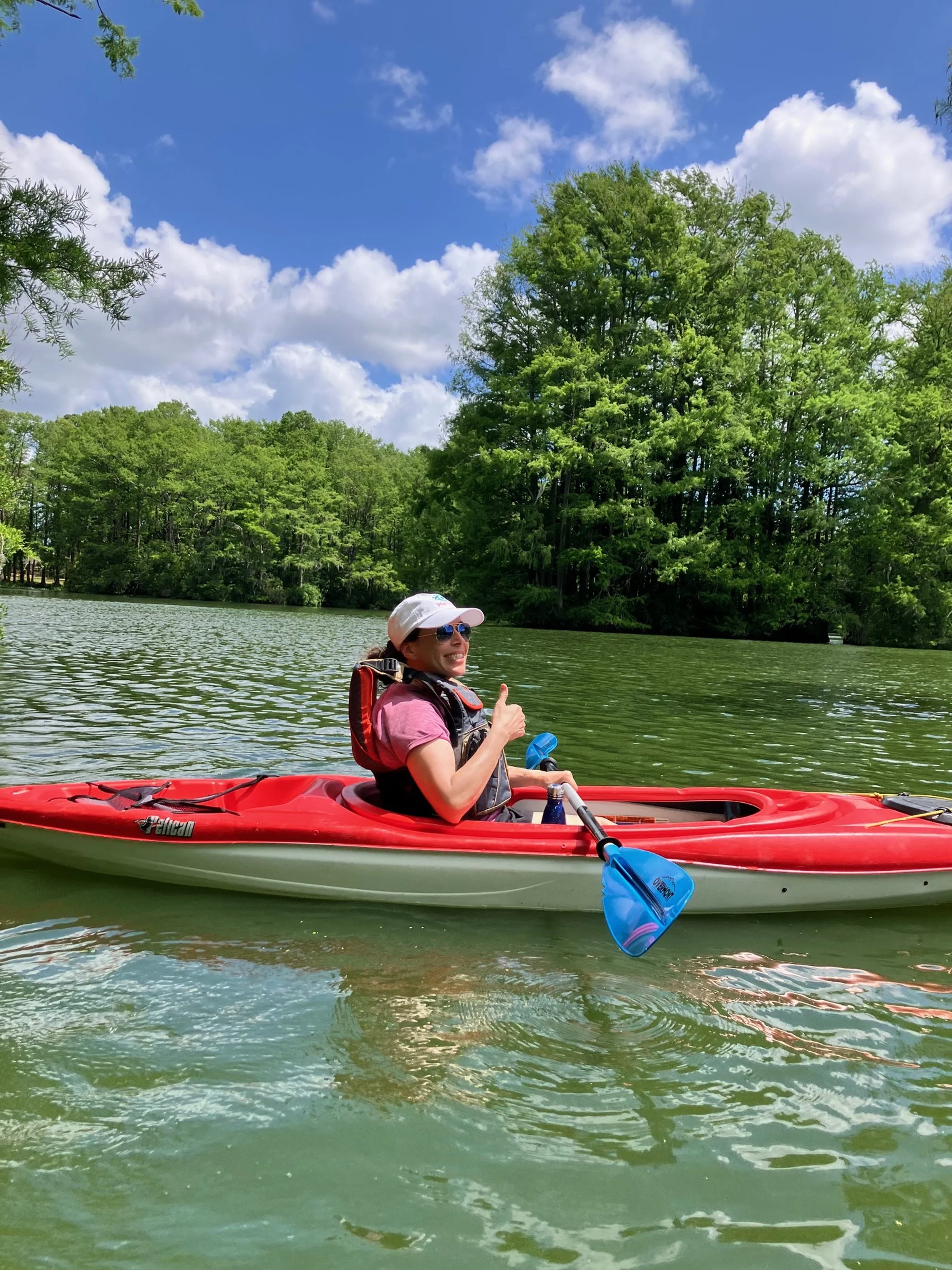 Woman in sunglasses smiling and giving a thumbs-up while kayaking on a lake with a green shoreline and a blue sky with white clouds.