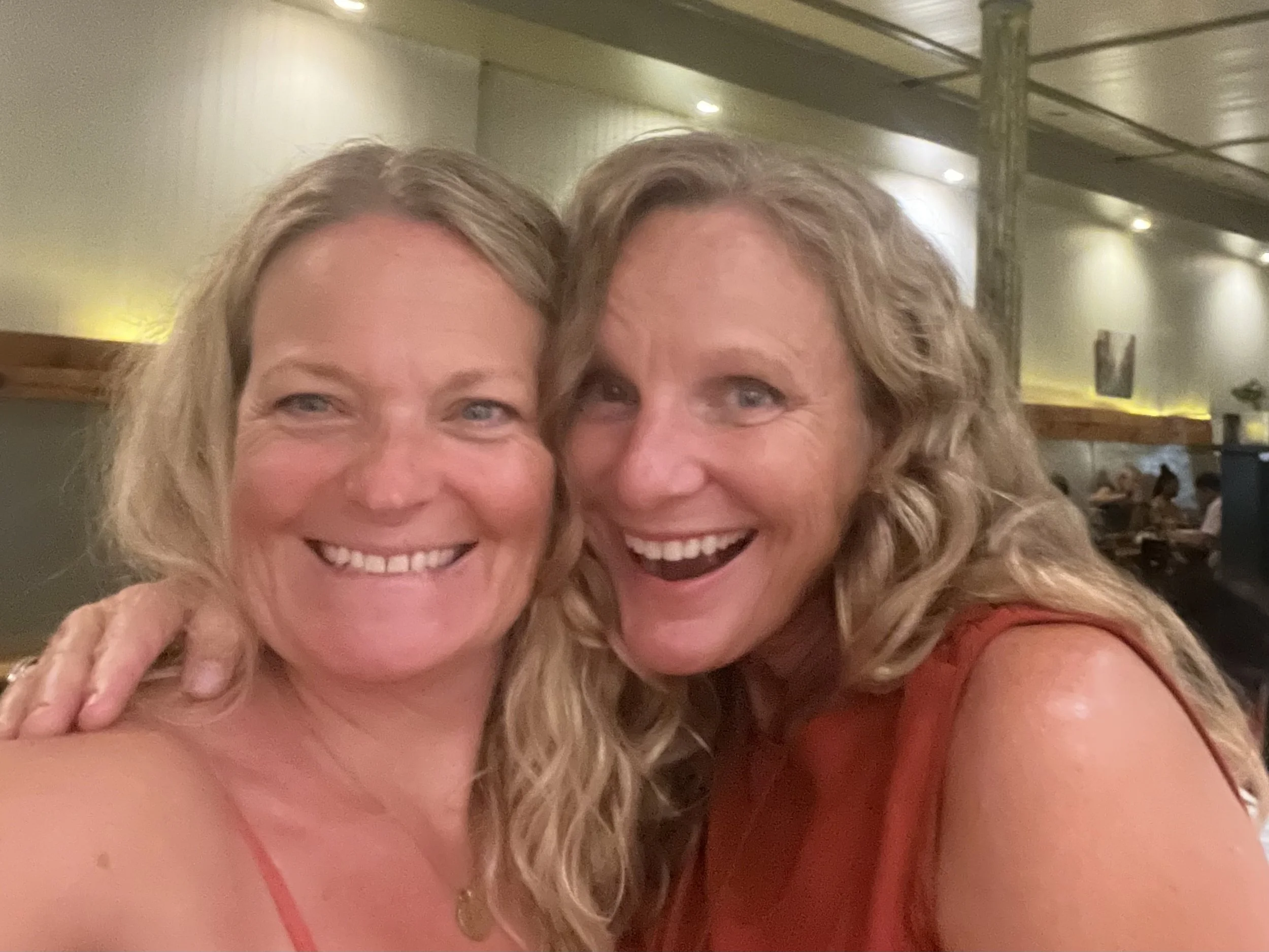 Two smiling women with curly hair taking a close-up selfie in a restaurant or bar.
