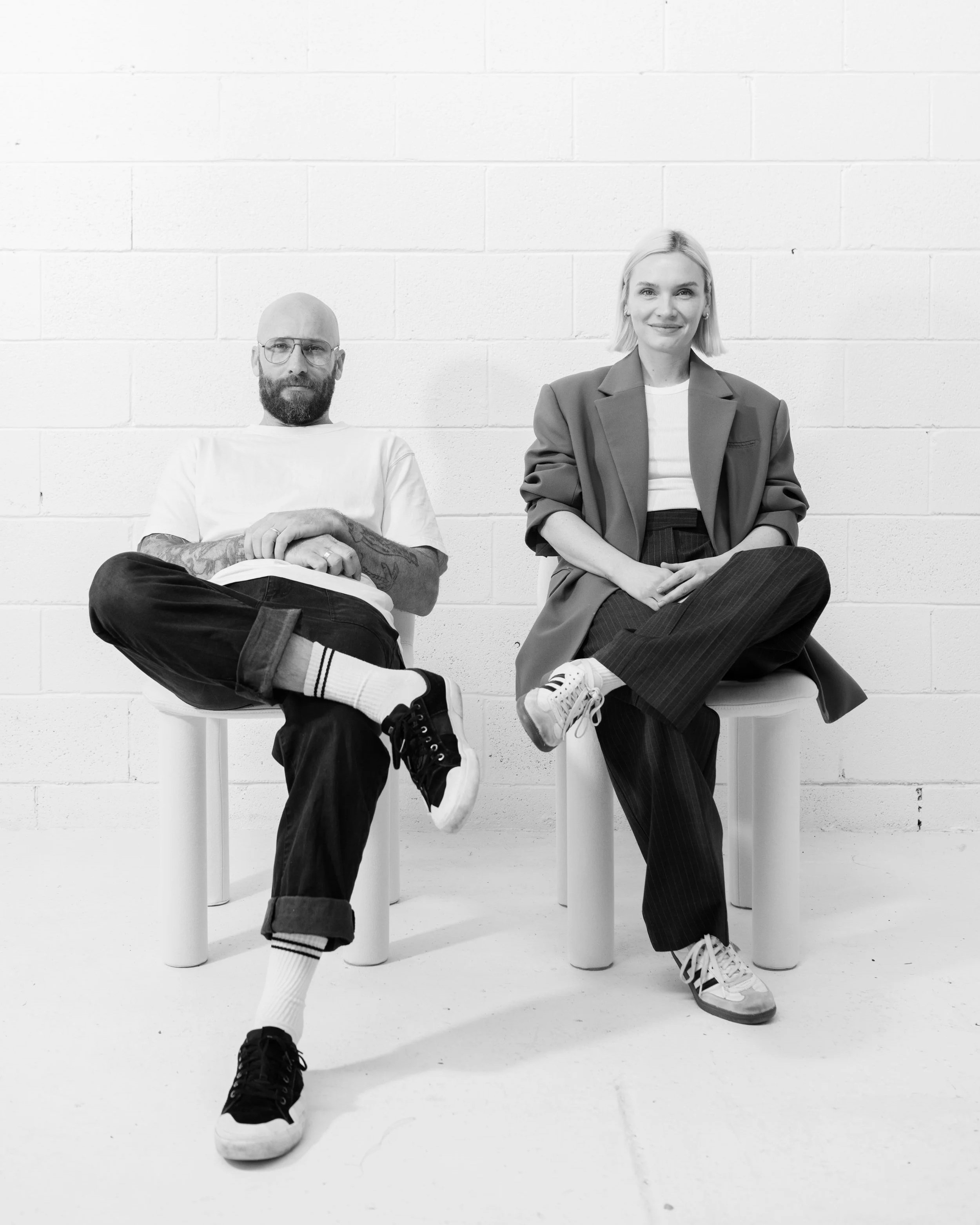 Black and white photo of a man and a woman sitting on stools against a plain white brick wall, smiling and looking at the camera. The man is bald with a beard, wearing glasses, a white shirt, tattoos on his arms, black pants, and black sneakers with white soles. The woman has short blonde hair, wearing a blazer over a white top, striped pants, and sneakers with laces. Their legs are crossed, and they appear relaxed and confident.