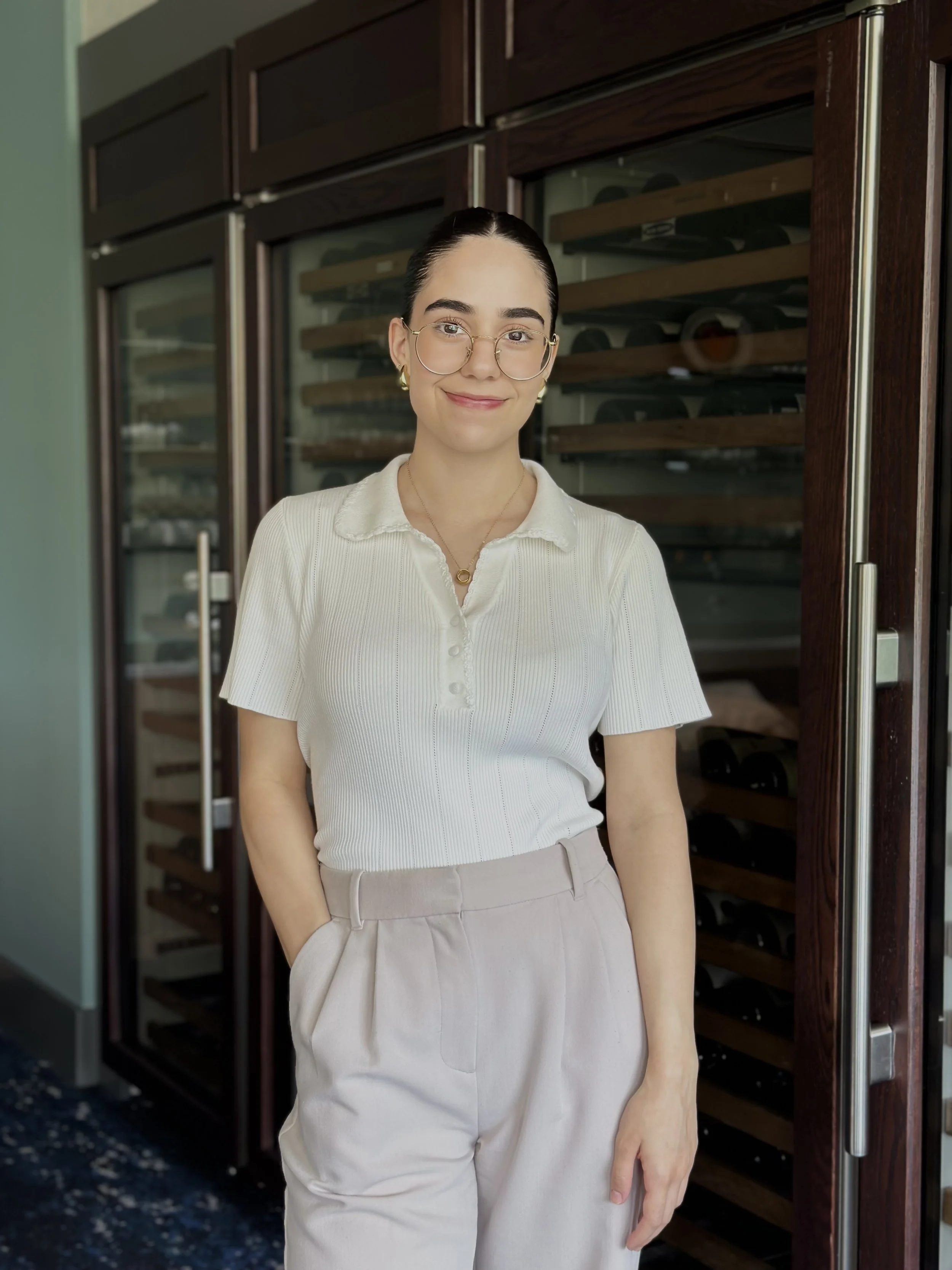 A smiling woman standing in a restaurant with wooden tables and large windows in the background, wearing a floral blouse with yellow and brown flowers.