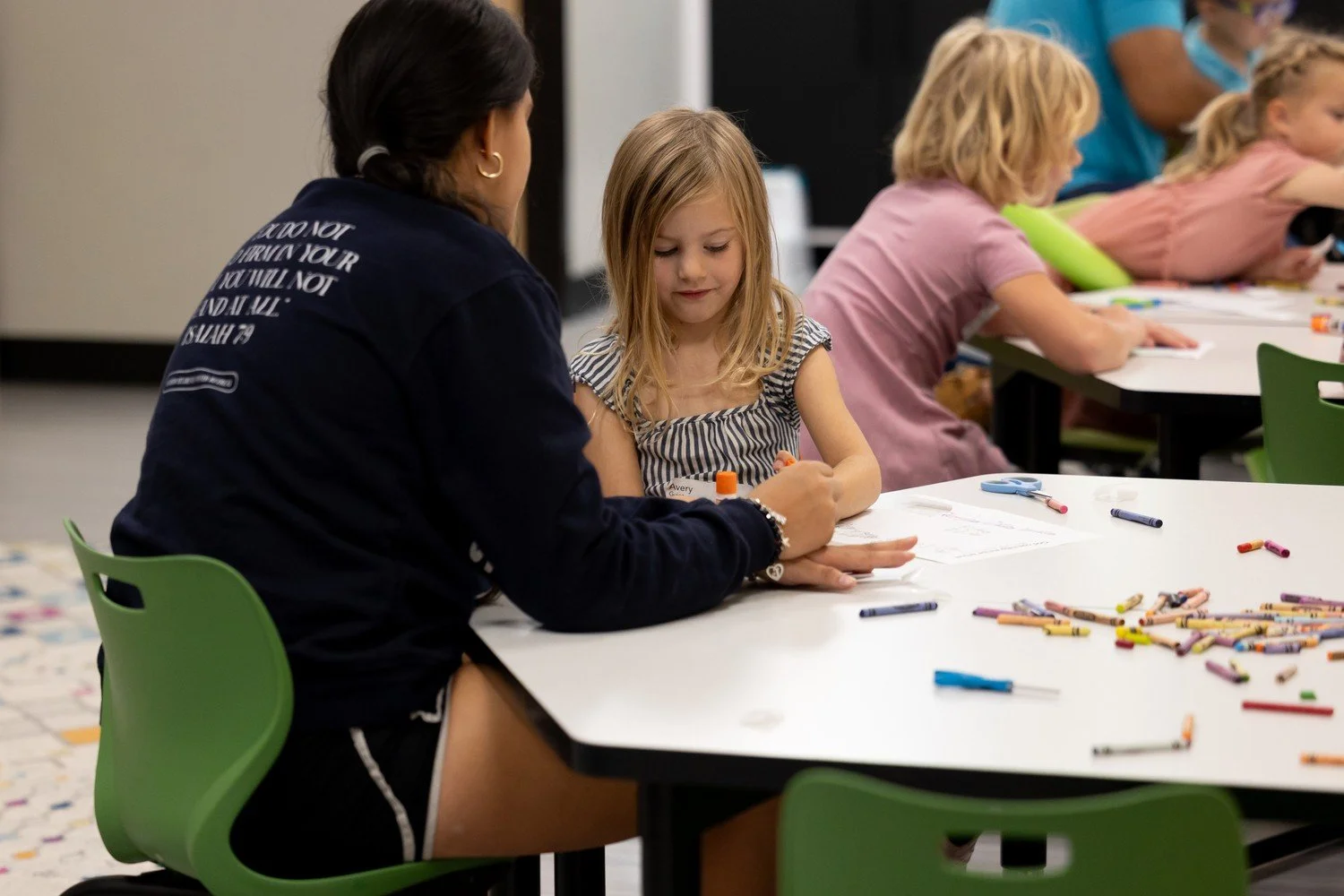 Children and teacher working on crafts at a classroom table, with crayons and paper.