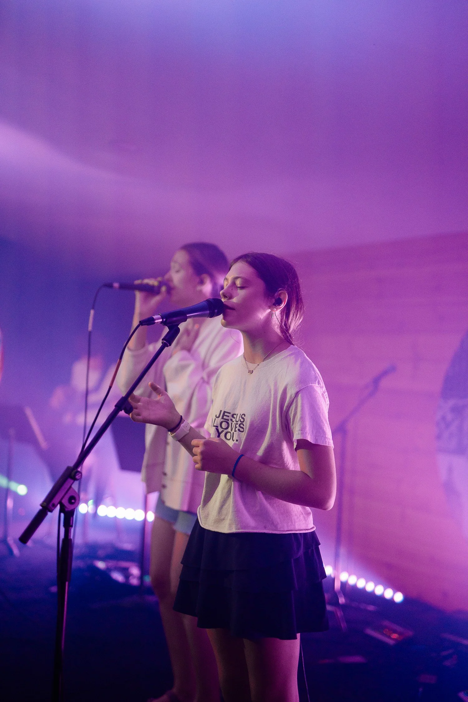Two women singing on stage with microphones, wearing casual clothing, with purple lighting.