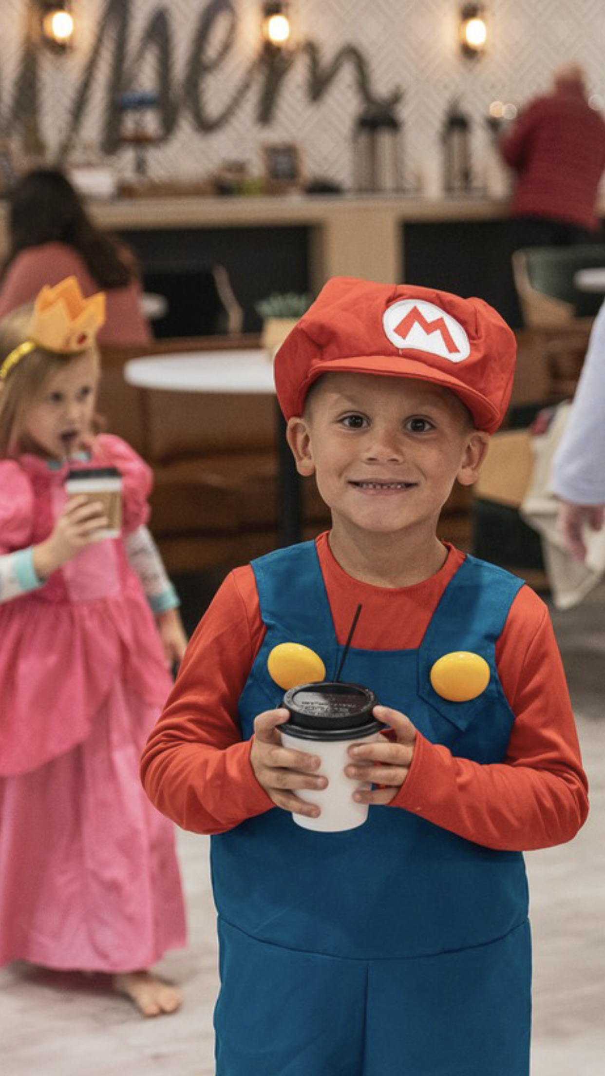 Children dressed in costumes holding cups; one as a red-capped character, another in a pink dress, standing indoors.
