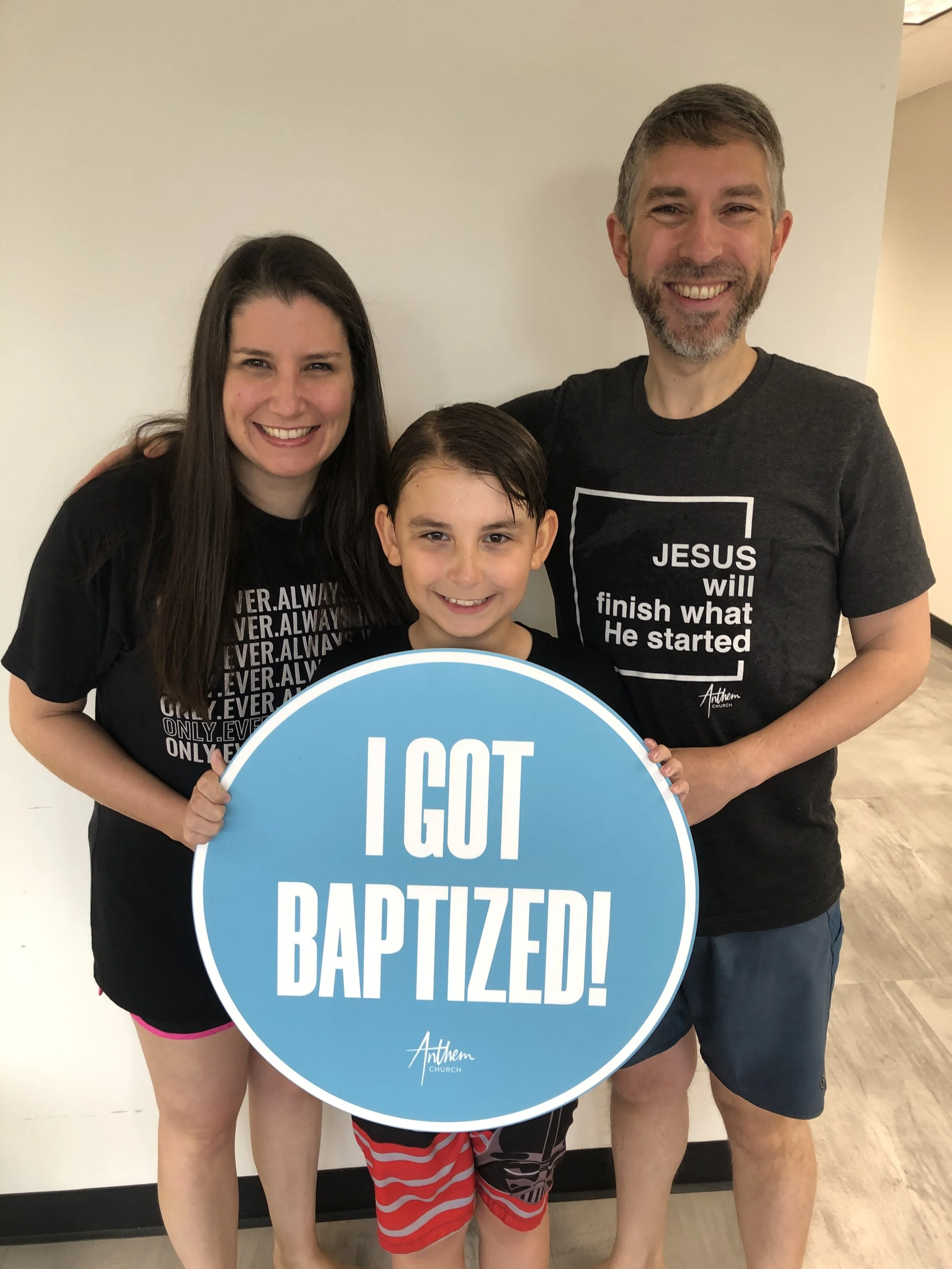 Three people smiling, including a child holding a sign that reads "I GOT BAPTIZED!"