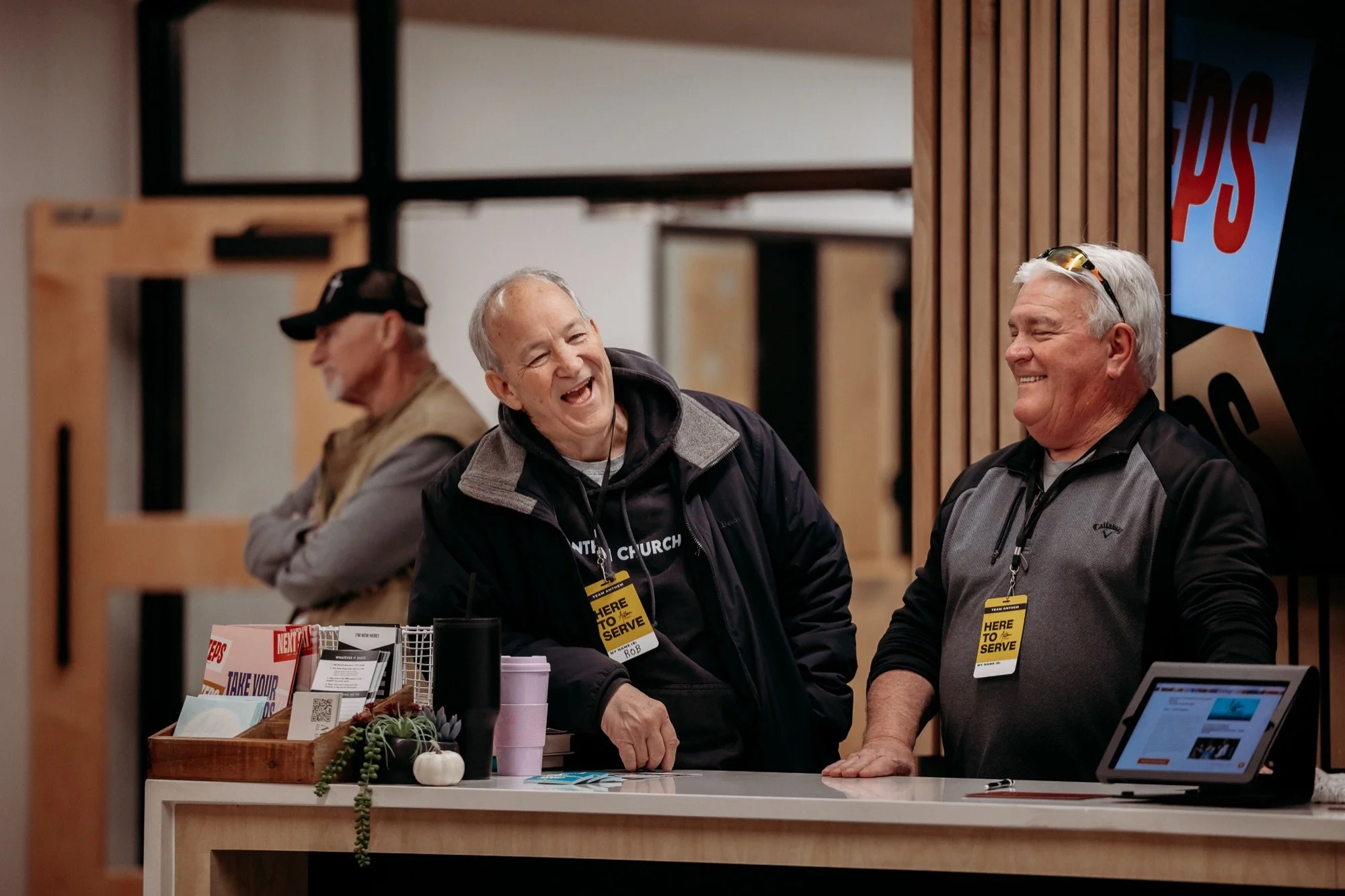 Three men smiling and talking at a recognition event, wearing lanyards and name tags that say 'HERE TO SERVE.' They are standing behind a table with brochures and small decorative items. The background features a wooden wall and a partial screen with bold text.