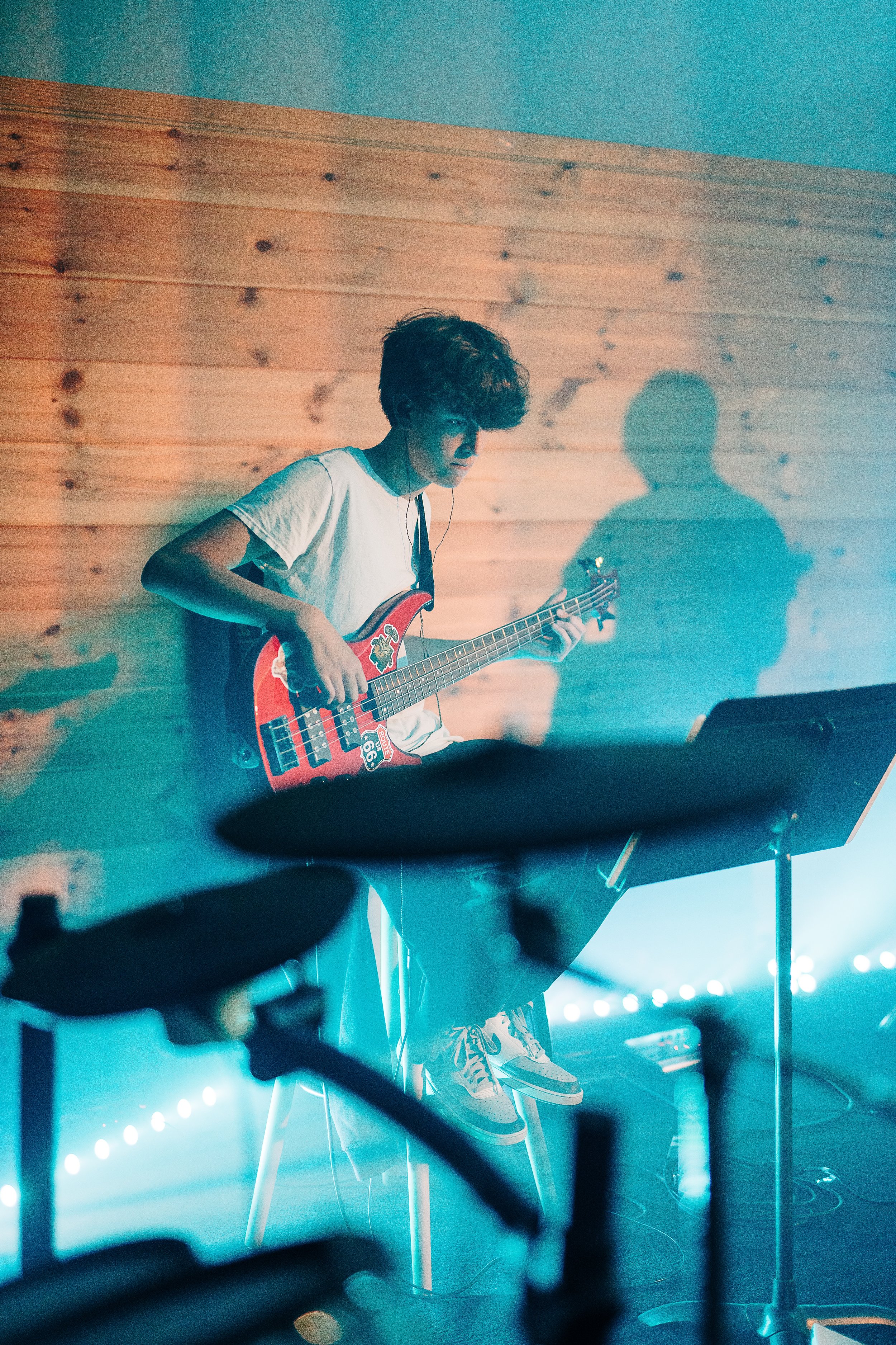 Person playing red electric bass guitar on stool in music studio, with drum set in foreground, wood-paneled wall, ambient blue lighting.