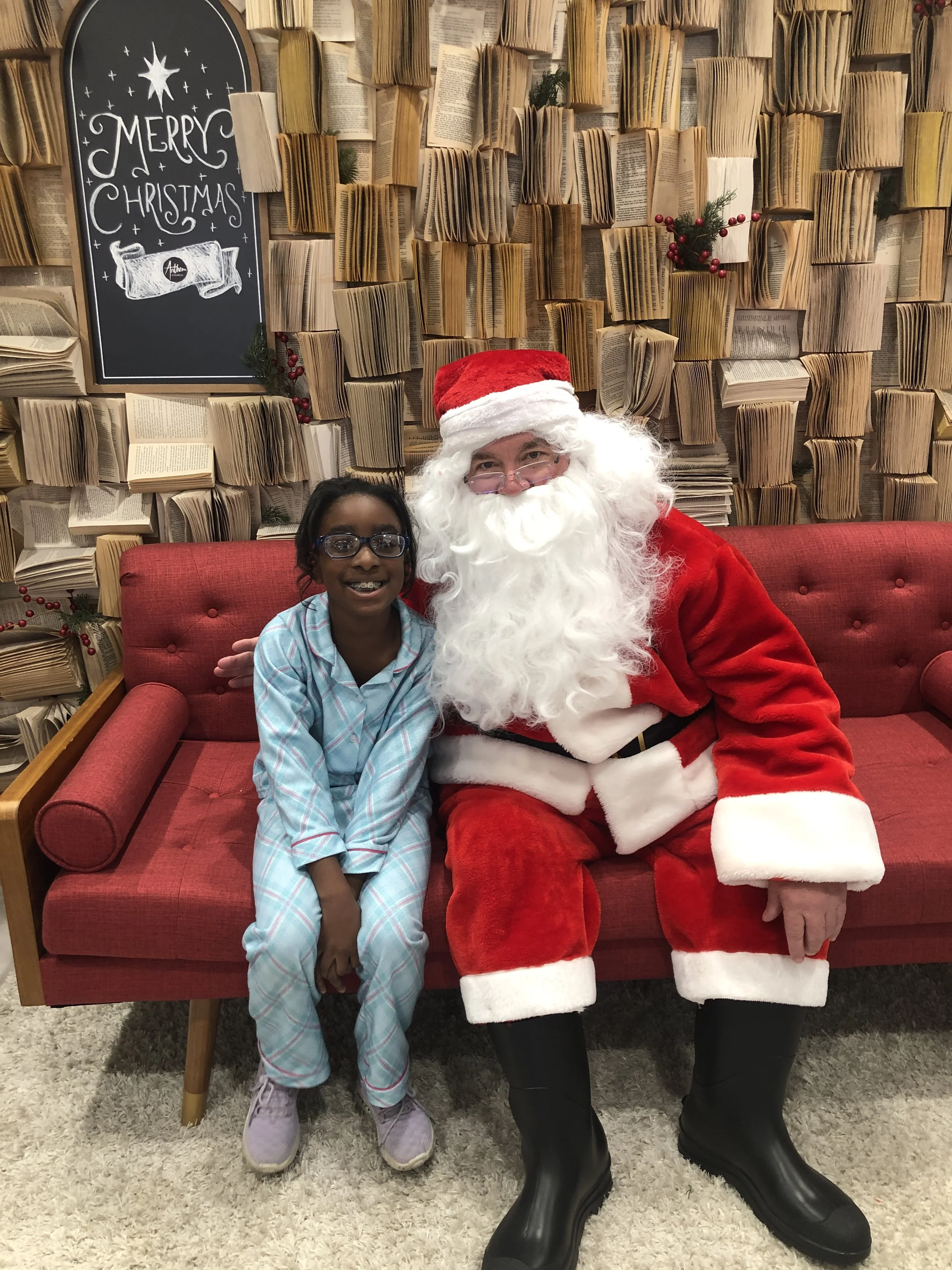 Child sitting with Santa Claus on a red couch, with a festive background and a sign saying "Merry Christmas."