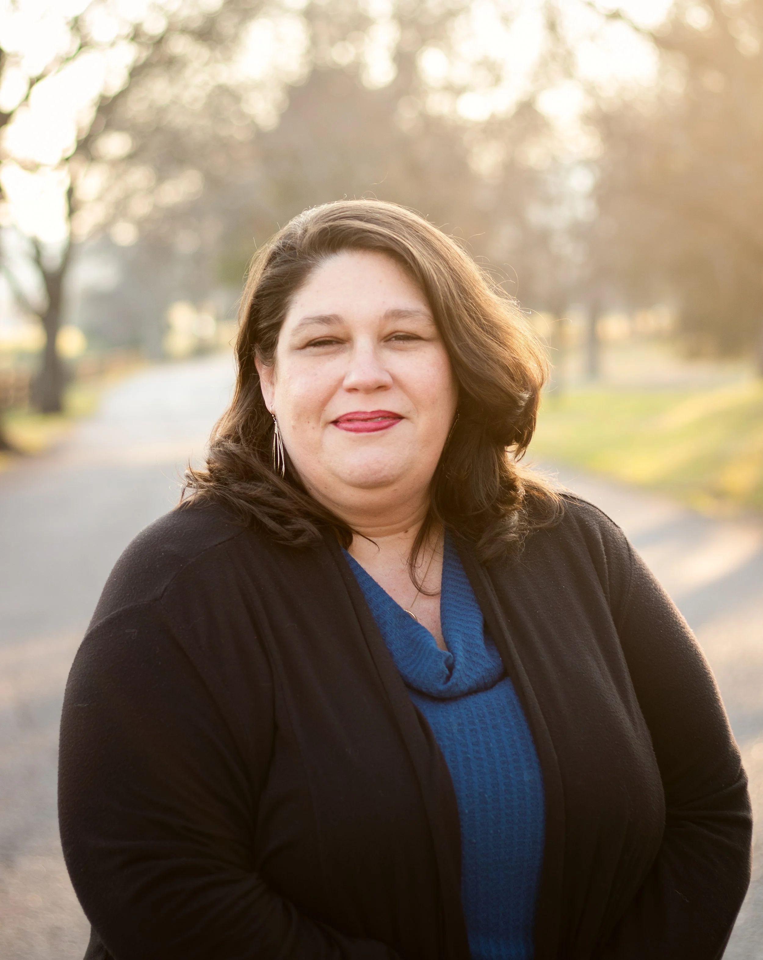 A woman with shoulder-length dark hair, wearing a black jacket and blue sweater, stands outdoors during golden hour with trees and a pathway in the background.