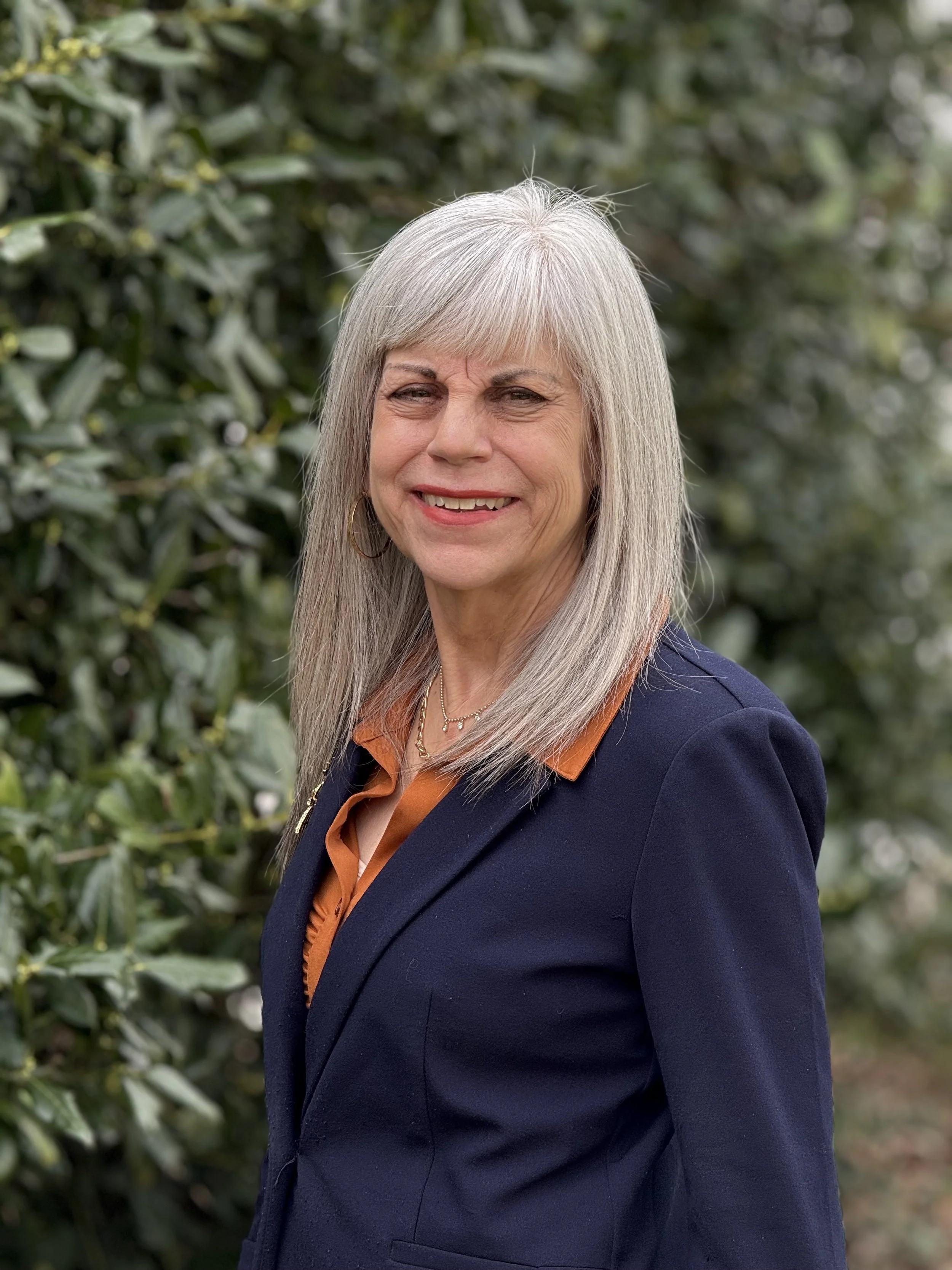 A smiling older woman with long gray hair, wearing a navy blazer over an orange shirt, standing outdoors with green foliage in the background.