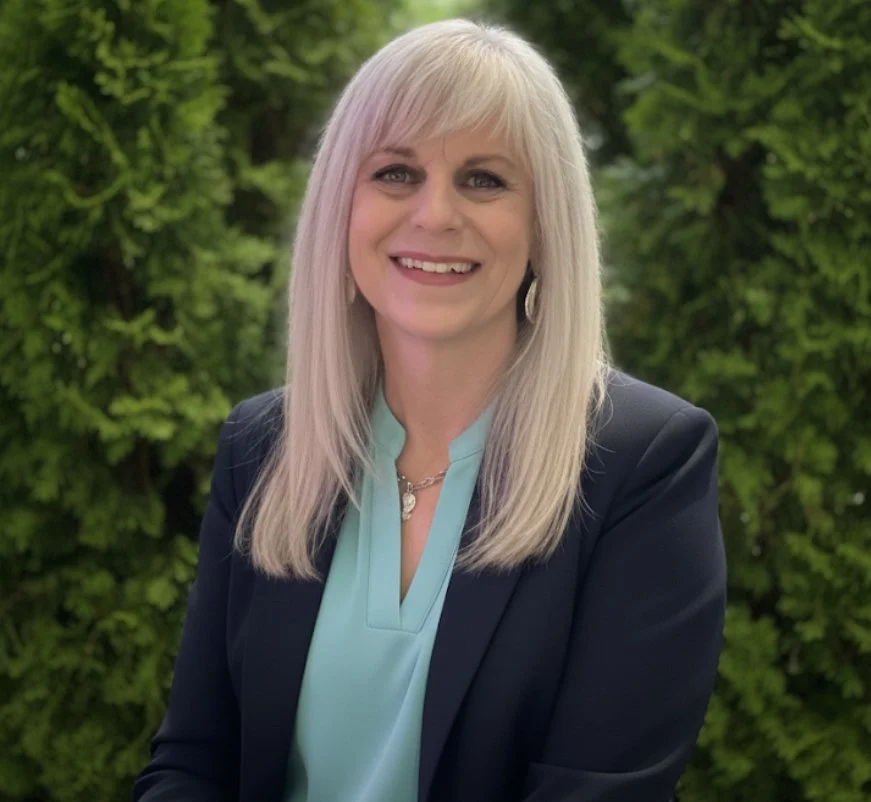 A smiling older woman with long gray hair, wearing a navy blazer over an orange shirt, standing outdoors with green foliage in the background.