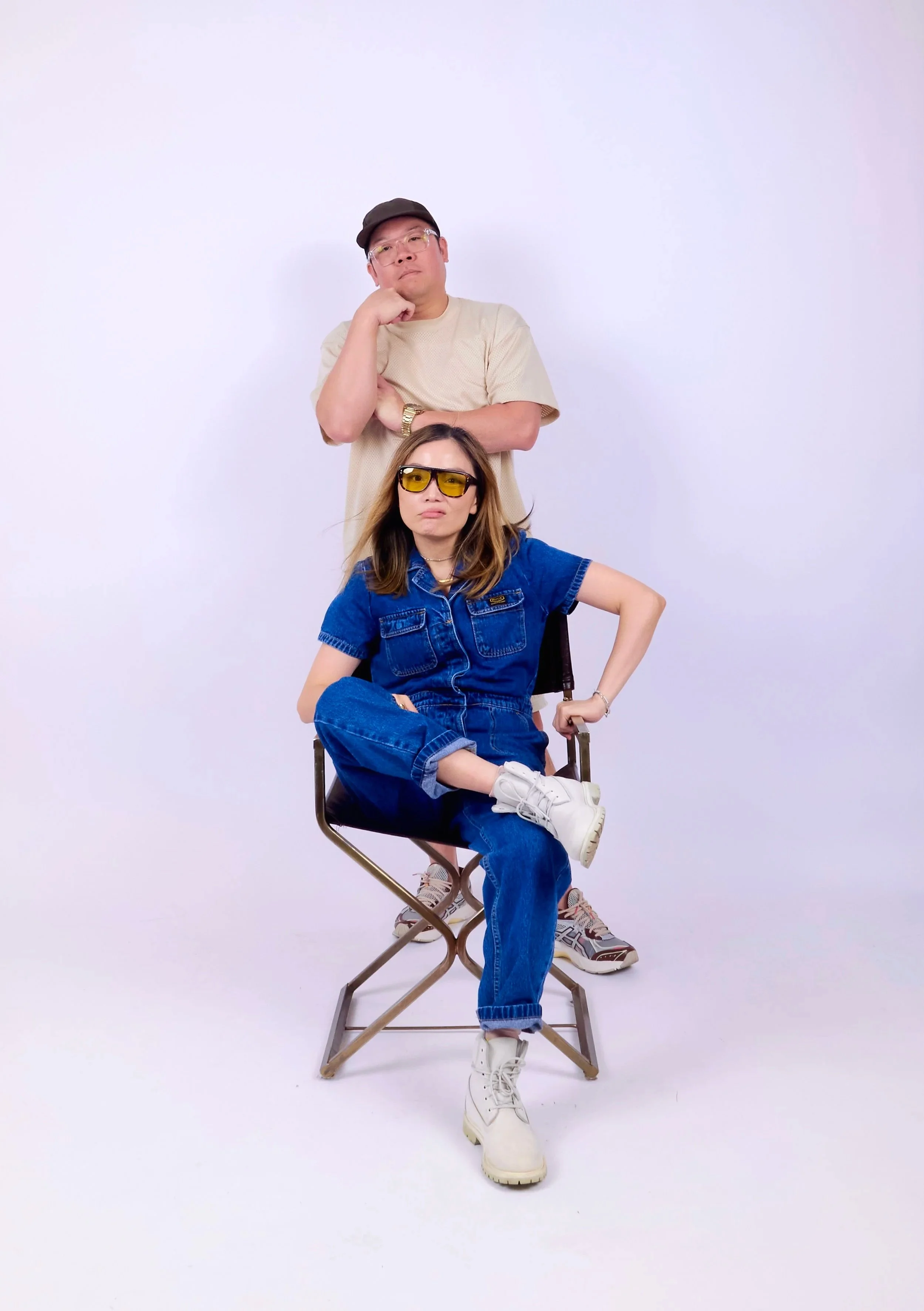 A man and woman posing in a photo studio with a plain white background. The man, standing behind the woman, is wearing glasses, a beige t-shirt, a brown cap, and a gold watch, with one hand resting on his chin. The woman, sitting on a chair, is weari