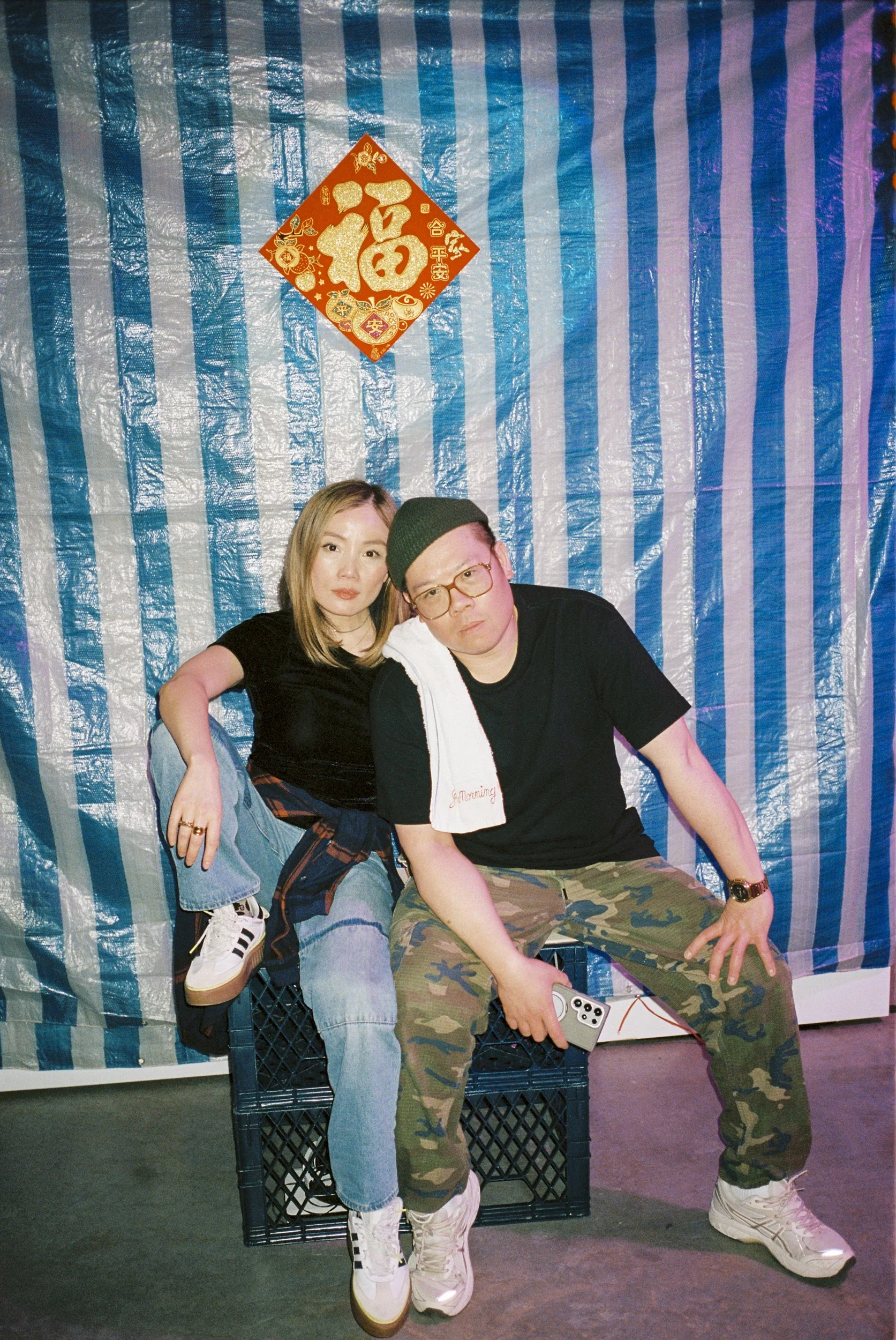 A man and woman sitting on a black crate in front of a shiny, blue and purple striped backdrop with a red Chinese decoration above them. The woman is wearing a black shirt, blue jeans, and white sneakers, while the man is dressed in a black shirt, ca