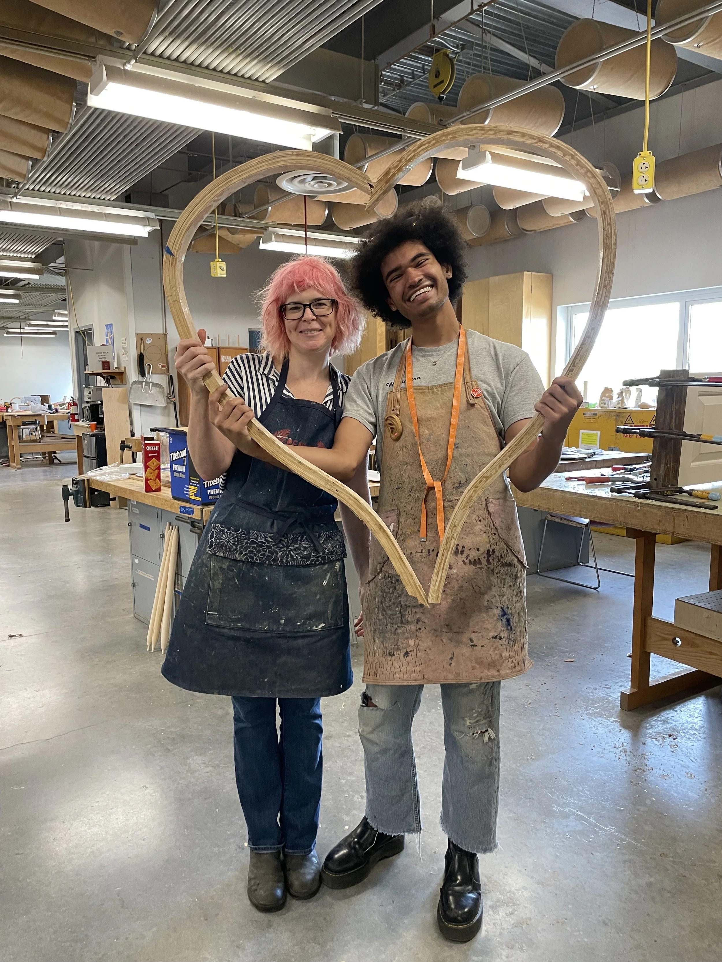 Woodworking instructor Annie Evelyn stands with student Justin R Cockrell in VCU's woodshop. The two are framed by a large heart made out of wood.