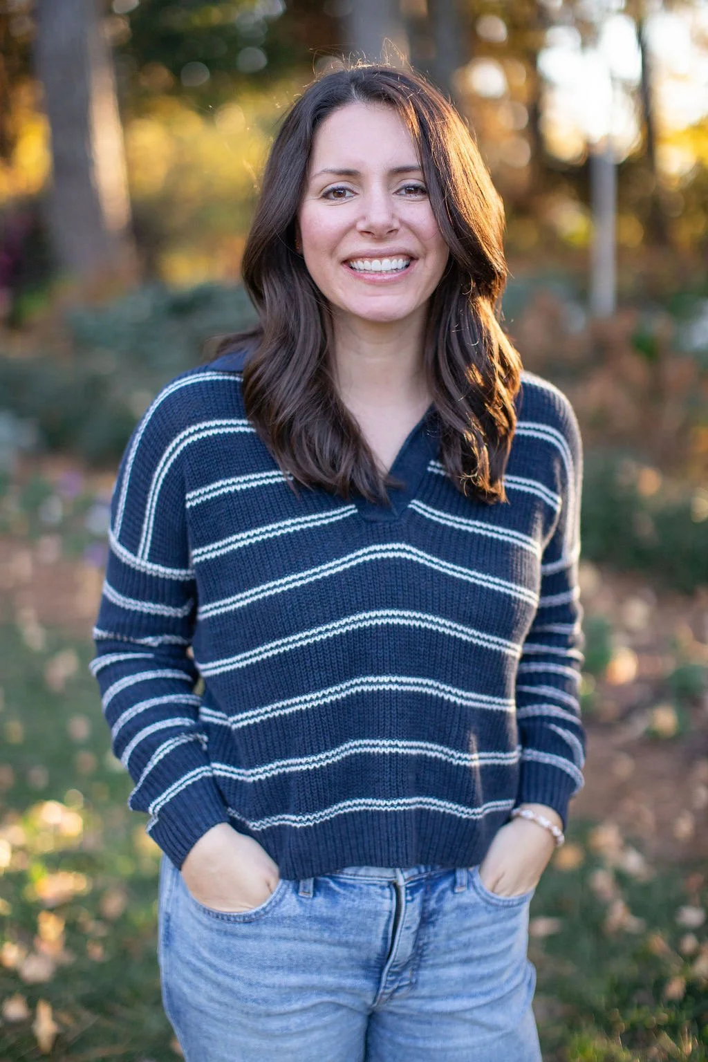 A woman with long dark hair, wearing a blue and white striped sweater and blue jeans, smiling outdoors during daytime with trees and sunlight in the background.