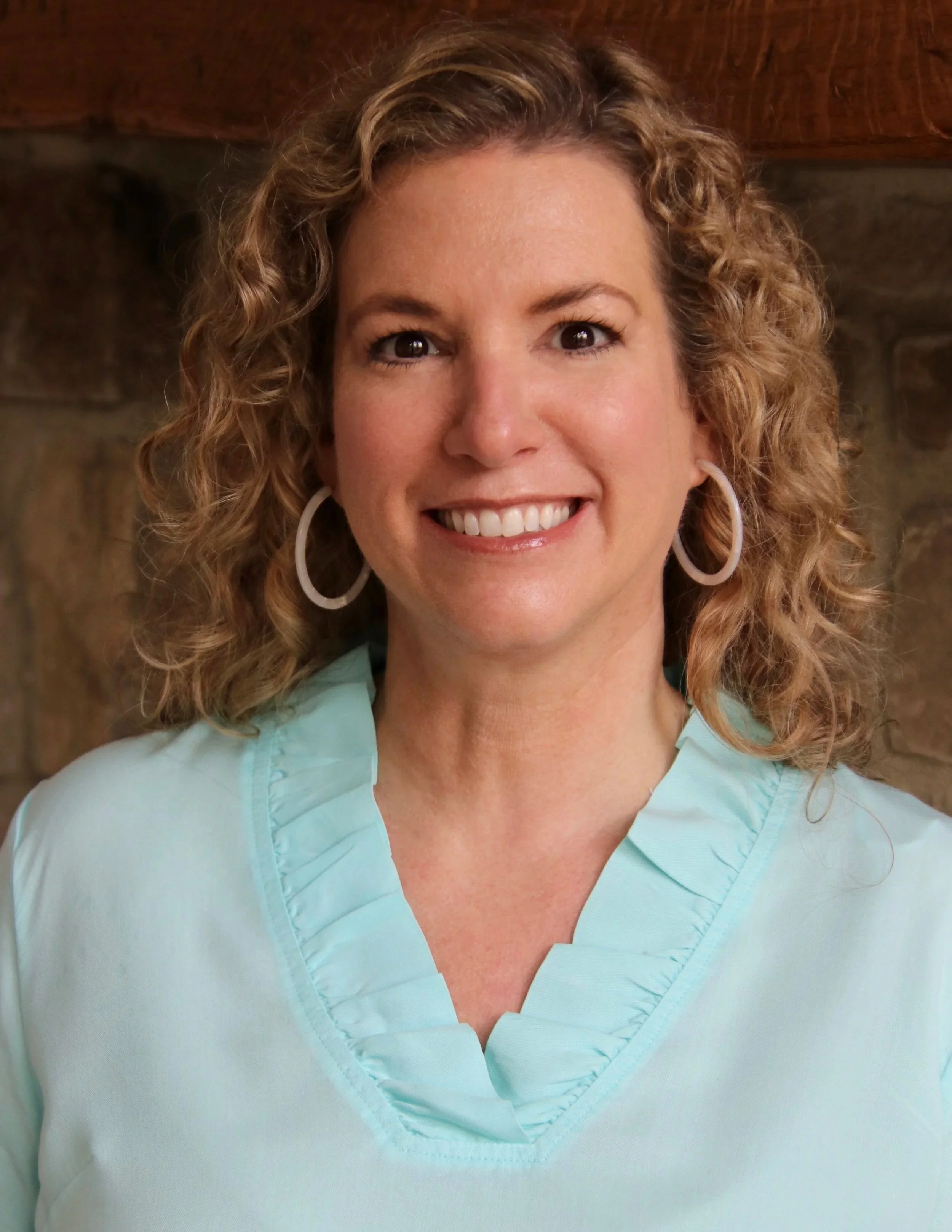 Close-up of a woman smiling, wearing a light turquoise medical scrub top and large white hoop earrings, with curly light brown hair, in front of a wooden and stone background.