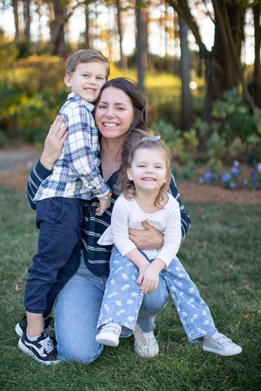 A woman with two young children outdoors in a wooded area, smiling and hugging. The boy is standing next to her, and the girl is sitting on her lap. All are smiling and appear happy.