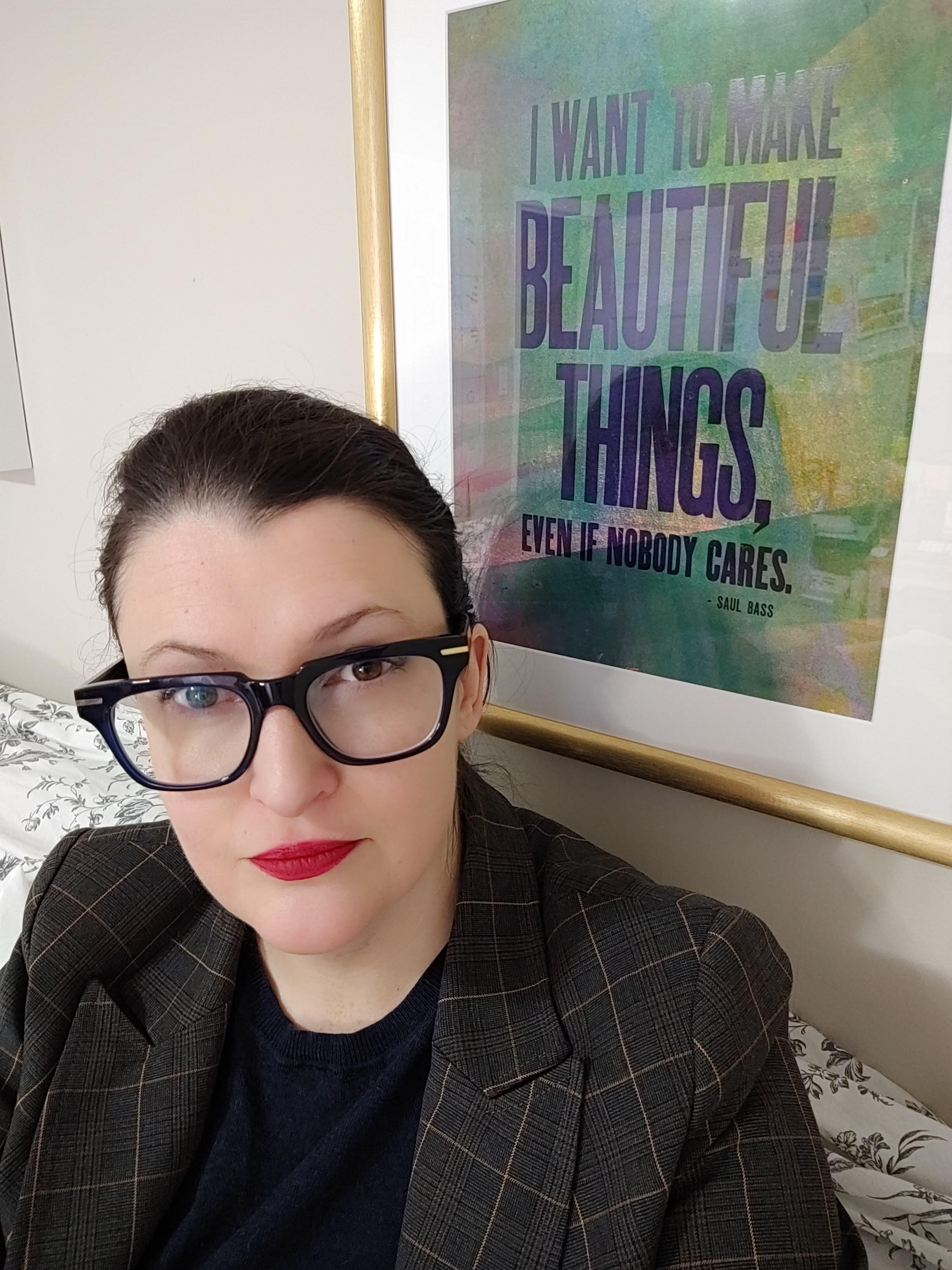 A person wearing thick framed glasses and a blazer, looking into the camera. They are sitting in front of an art print that reads "I want to make beautiful things, even if nobody cares."
