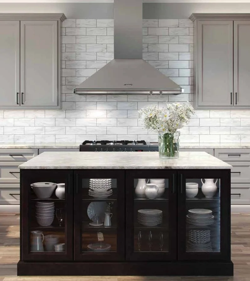 Modern kitchen featuring grey cabinets, a marble countertop island with glass-front cabinets filled with white dishes, a stove with a stainless steel range hood, a white marble backsplash, and a vase of white flowers on the island.