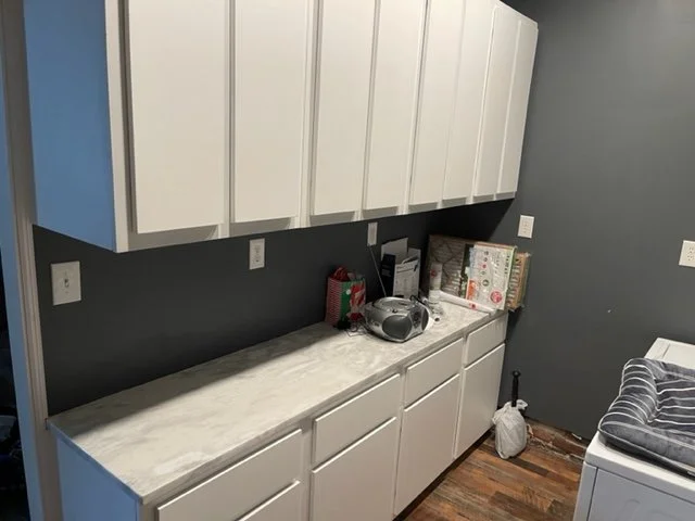 Kitchen counter with white cabinets above and below, a gray wall, and various items including a radio, books, and a basket. Part of a washing machine is visible on the right side.