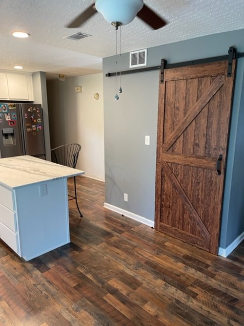 Interior of a home with a gray wall, a wooden sliding barn door, a ceiling fan, a refrigerator, and a kitchen island with a black chair.