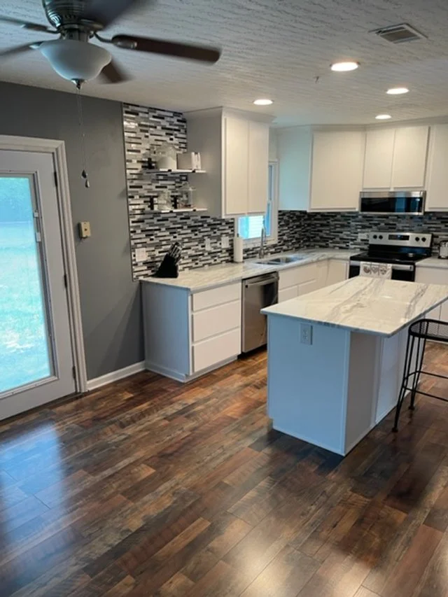 Modern kitchen with white cabinets, a marble island, stainless steel appliances, a gray and black backsplash, and wooden flooring.