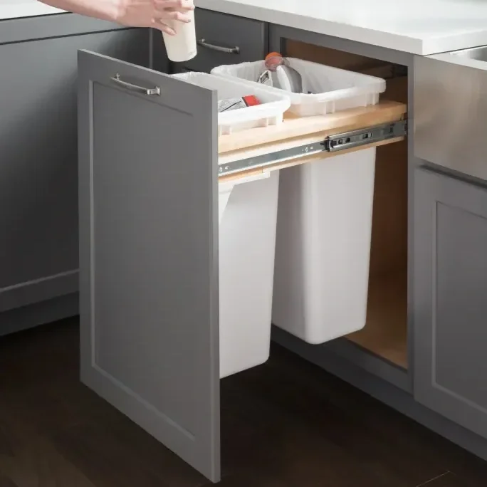 Open kitchen cabinet with trash and recycling bins, with a person's hand holding a cup above the bins.