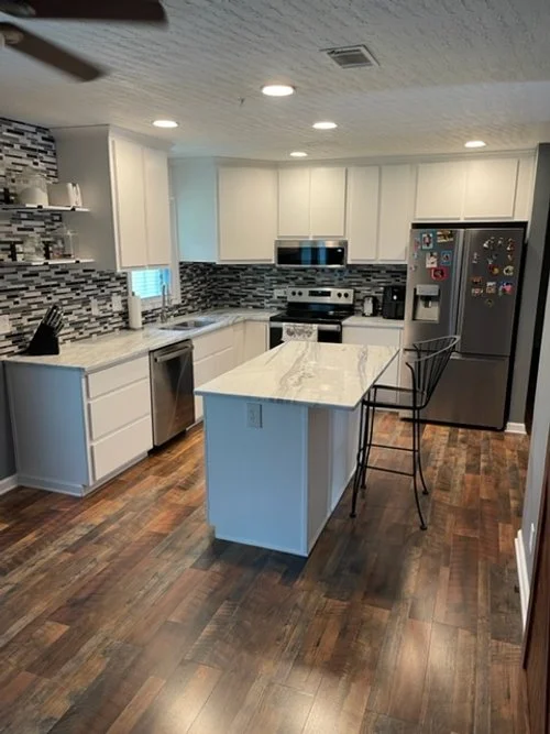 Modern kitchen with white cabinets, stainless steel appliances, a marble countertop island, black bar stools, and hardwood flooring.